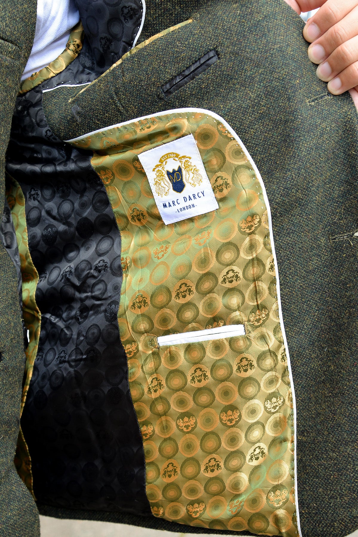 A close-up of the MARLOW Olive Green Tweed Blazer by marcdarcy shows its gold and green patterned lining and an inner pocket with a "marcdarcy" label. A hand holds open the slim fit men's wool-poly blend jacket.