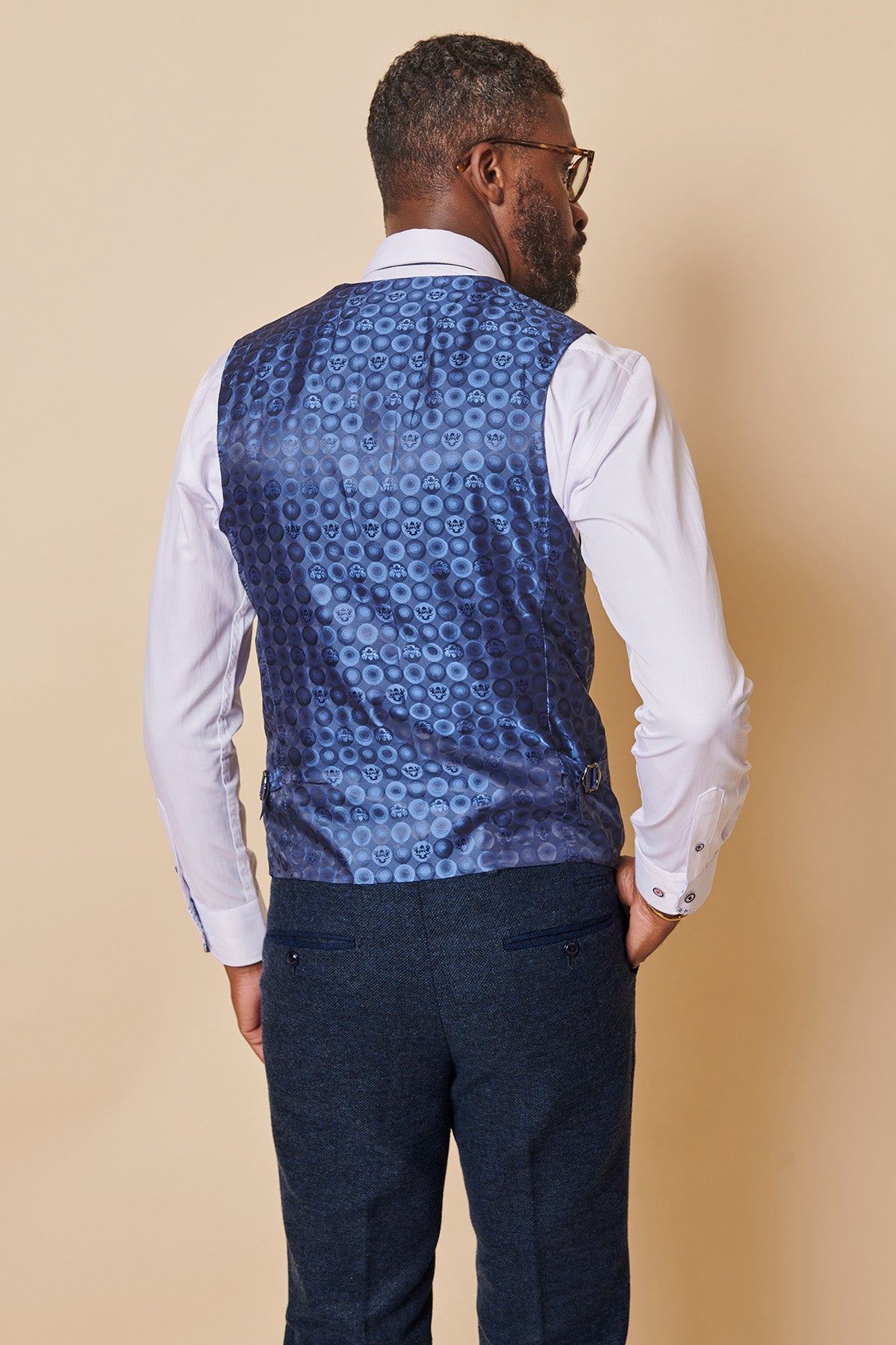 A man with short hair and glasses, wearing a slim fit white shirt, MARLOW - Navy Tweed Single Breasted Waistcoat by marcdarcy, and dark blue trousers stands facing away against a beige background.