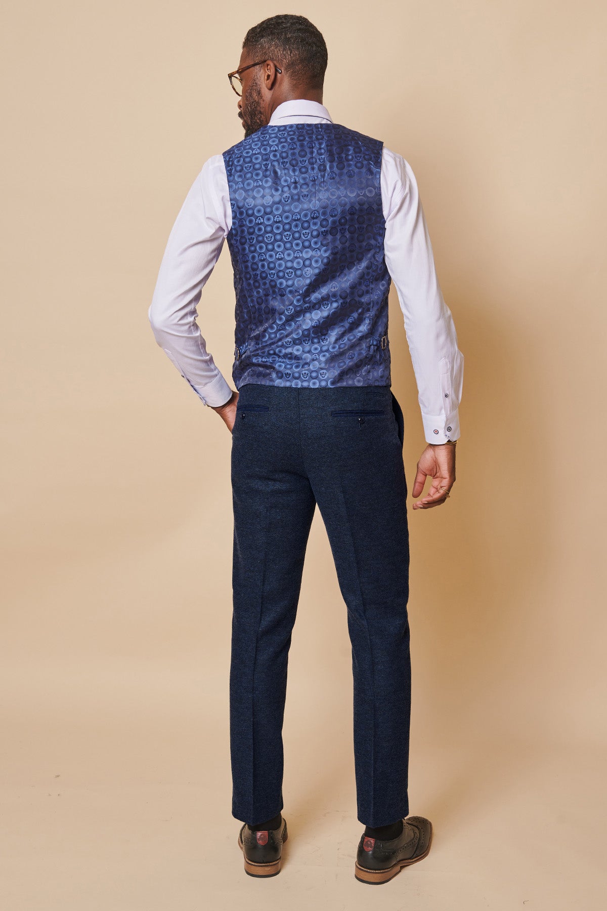 A man stands facing away, wearing a white shirt, the marcdarcy MARLOW navy tweed single breasted waistcoat, dark blue trousers, and brown shoes against a plain beige background.