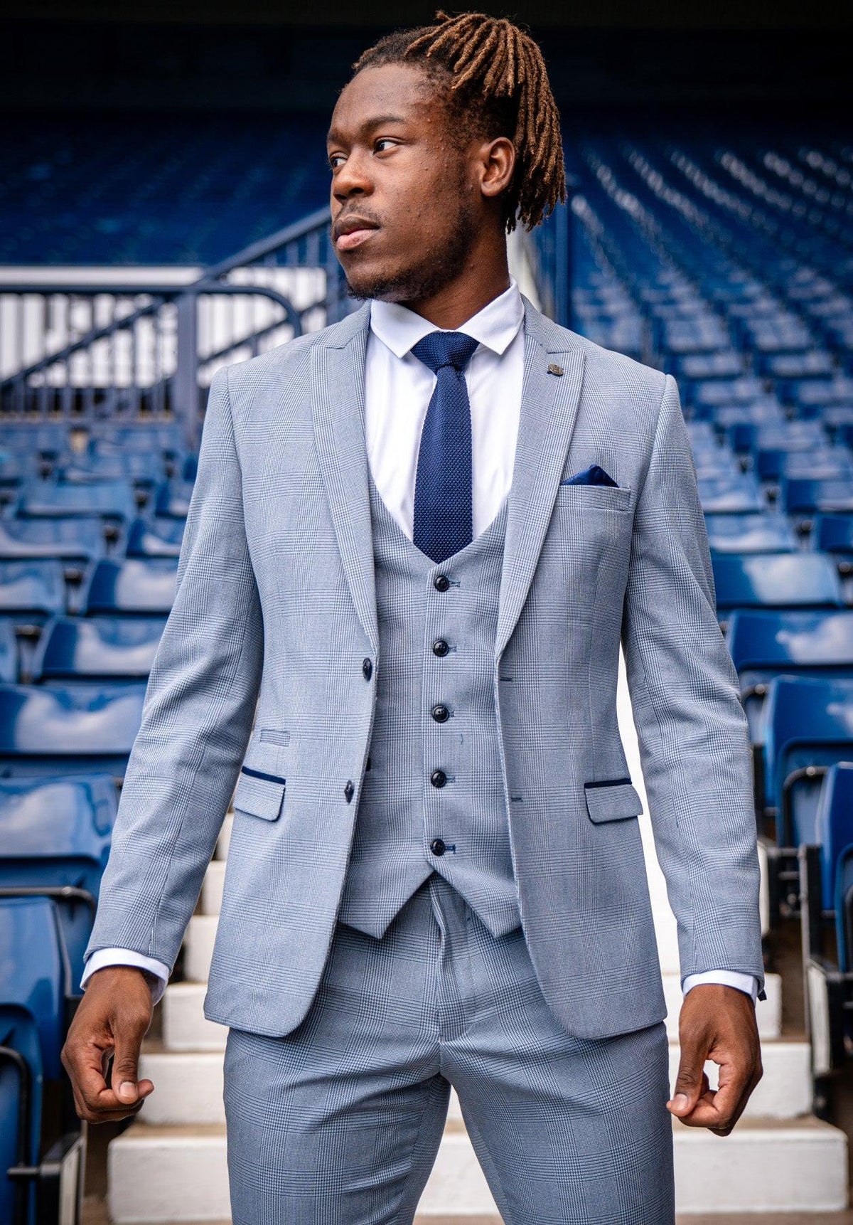 A man stands confidently in stadium seating, wearing the Marc Darcy WBA Collection - BROMLEY Suit as worn by Brandon Thomas-Asante. The light gray Prince of Wales check contrasts with his navy tie, with empty seats behind him.