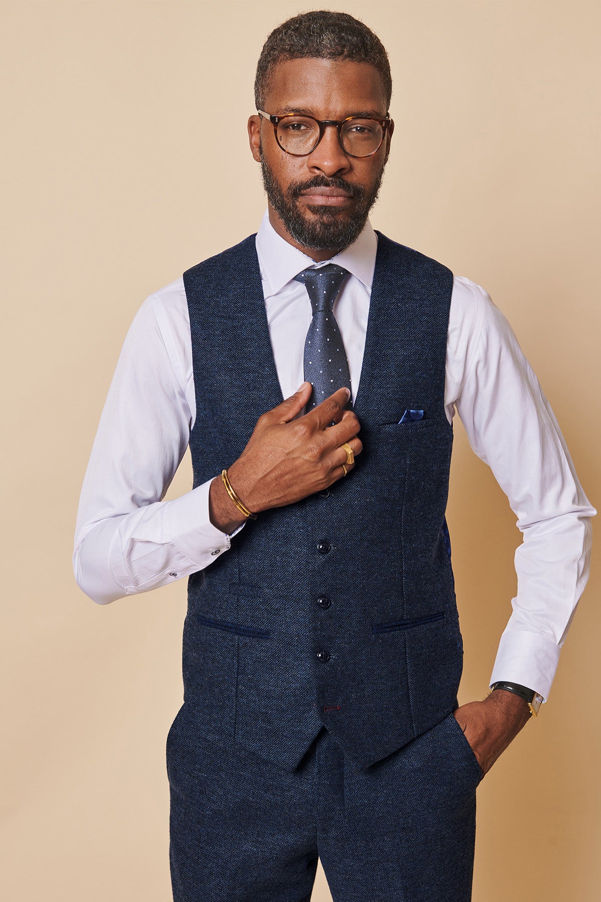 A man in glasses, a white shirt, blue polka-dot tie, and the marcdarcy MARLOW Navy Tweed Single Breasted Waistcoat (slim fit) stands against a beige background, looking at the camera while adjusting his vest.