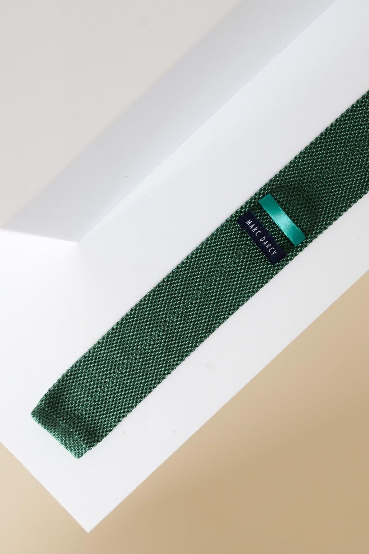 The Accessory KT - Olive Knitted Tie & Pocket Square features an olive green knit tie with a modern texture and navy Marc O’Polo tag, laid diagonally on a white geometric surface with a beige background.