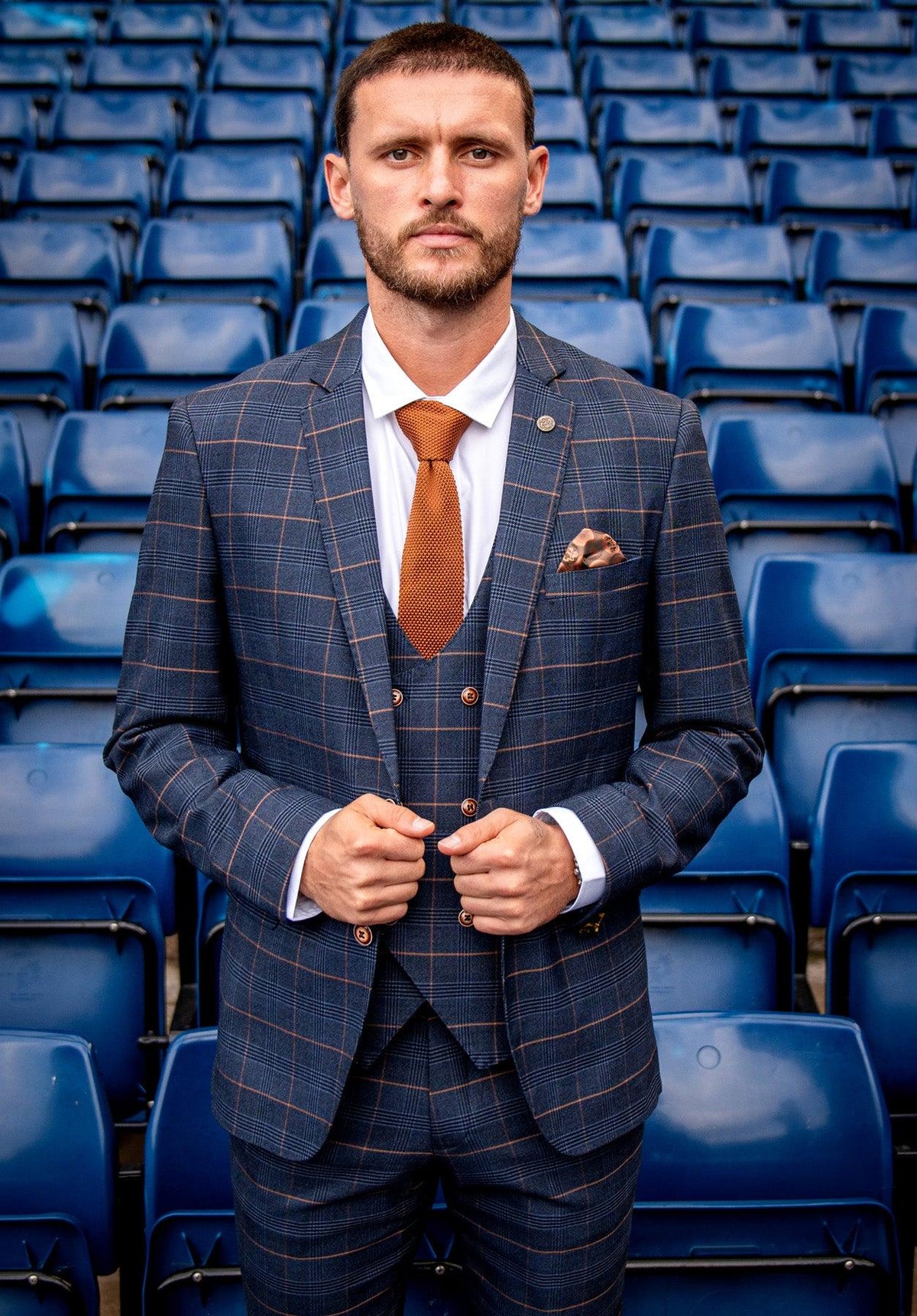 A man wears the marcdarcy WBA Collection - JENSON Marine Navy Check Suit as worn by John Swift, styled with an orange tie and pocket square, standing in front of blue stadium seats.
