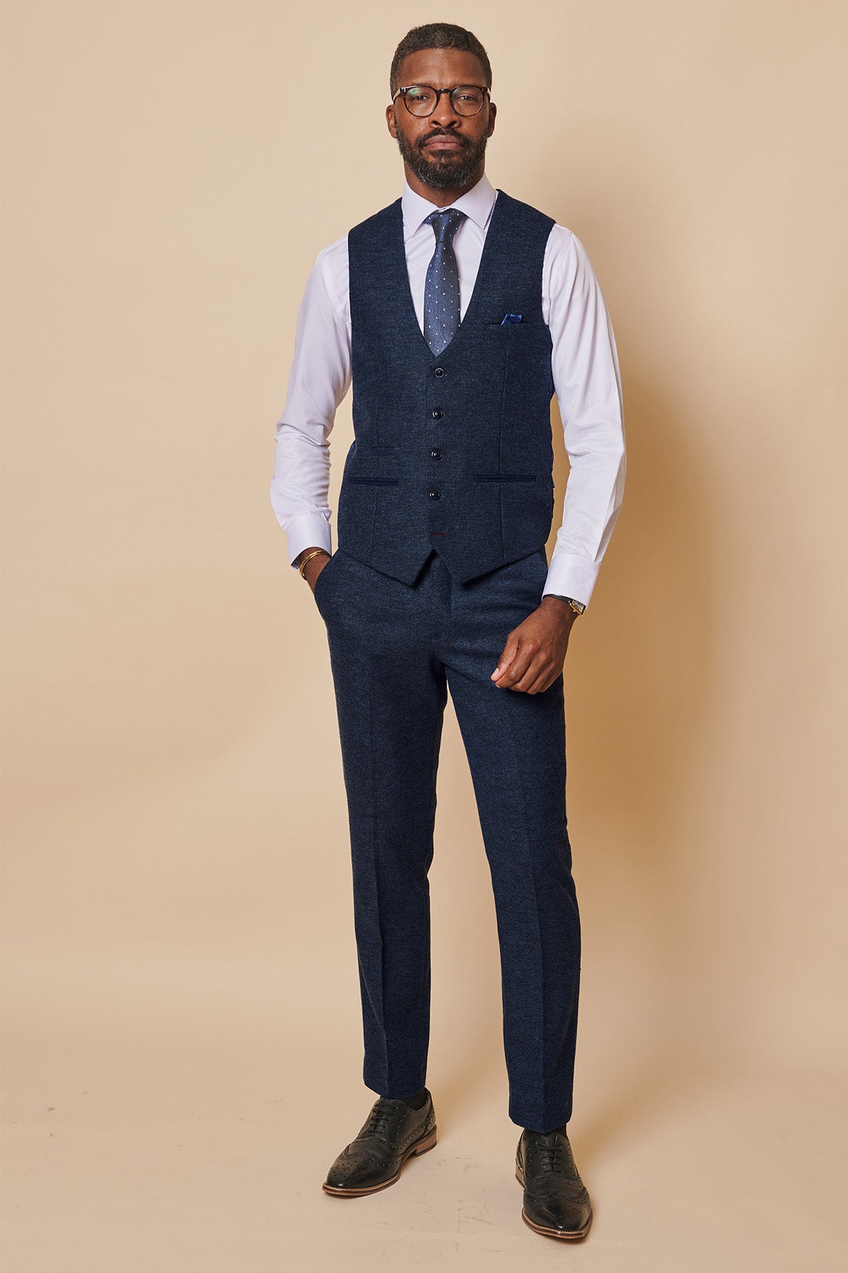 A man wearing glasses stands against a beige background in the marcdarcy MARLOW navy tweed single breasted waistcoat and matching trousers, paired with a white dress shirt, blue patterned tie, and black dress shoes, hands in his pockets.