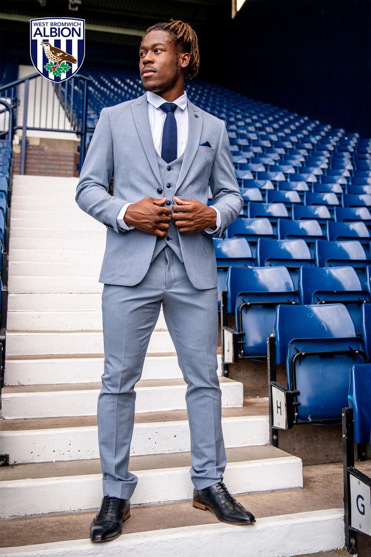 Brandon Thomas-Asante models the Marc Darcy BROMLEY suit from The WBA Collection, standing confidently by blue stadium seats with the West Bromwich Albion crest in view.