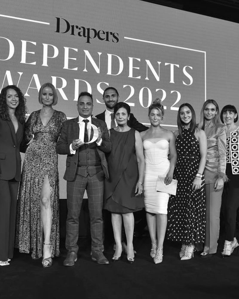 A group of people dressed formally stand on stage holding an award, smiling for a photo at the Drapers Independents Awards 2022, with the event name displayed on a large screen behind them.