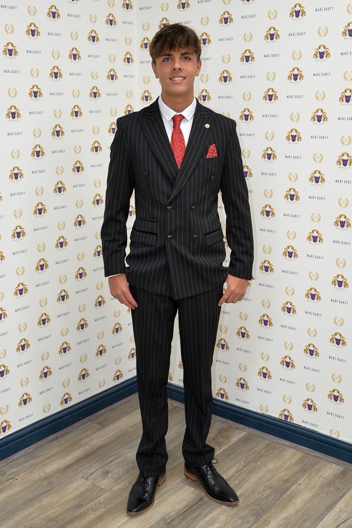 Luca Toolan as Mason Radcliffe smiles in front of a gold and purple logo backdrop, wearing the Marc Darcy ROCCO black pinstripe double-breasted two-piece suit from Coronation Street, paired with a red tie and white shirt on a wooden floor.