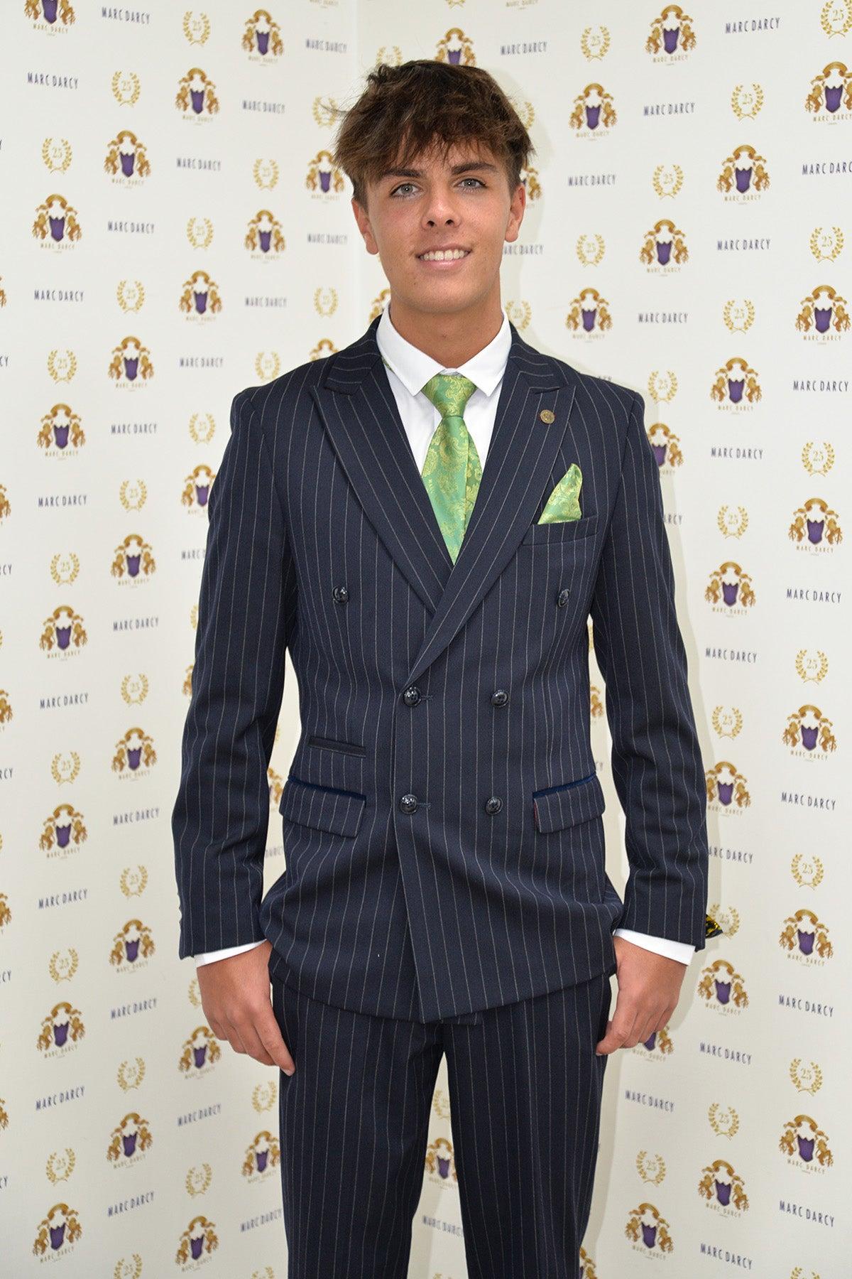 Coronation Street’s Mason Radcliffe (Luca Toolan) smiles in front of a Marc Darcy backdrop wearing the ROCCO Navy Pinstripe Double Breasted Two Piece Suit by Marc Darcy, paired with a white shirt and green tie.