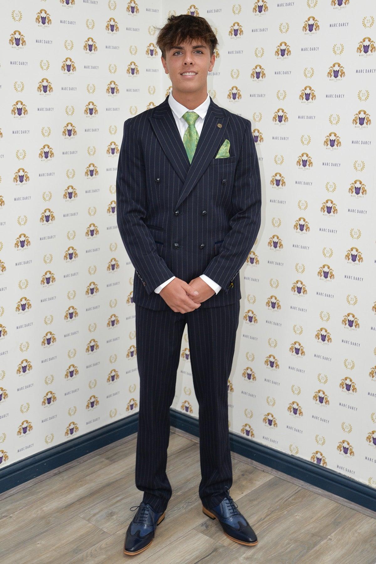 Coronation Street's Mason Radcliffe (Luca Toolan) smiles in front of a patterned backdrop, wearing the Marc Darcy ROCCO Navy Pinstripe Double Breasted Two Piece Suit with a green tie and pocket square, white shirt, and dress shoes.