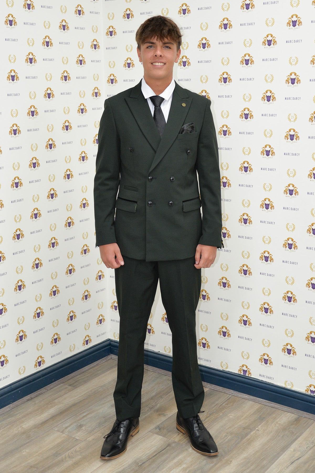 Coronation Street’s Mason Radcliffe (Luca Toolan) wears the Marc Darcy BROMLEY Olive Green Double Breasted Two Piece Suit, paired with a black tie, white shirt, and black oxford shoes, posing before a Marc Darcy branded backdrop.