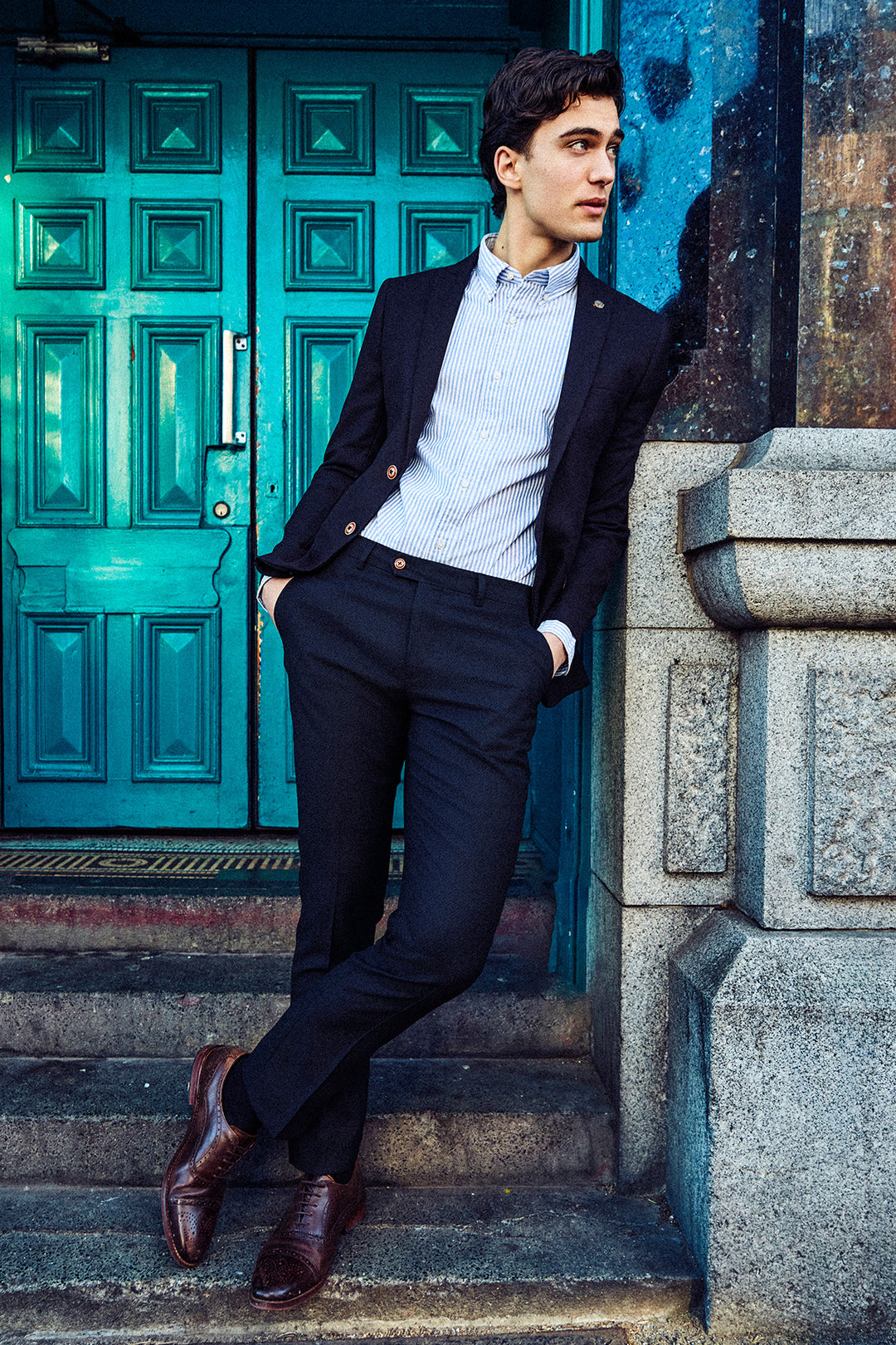 A man in the Marc Darcy MAX Navy Blue Two Piece Suit stands on stone steps, wearing a light striped shirt and brown dress shoes, leaning against a building with turquoise double doors, hands in pockets and looking to the side.