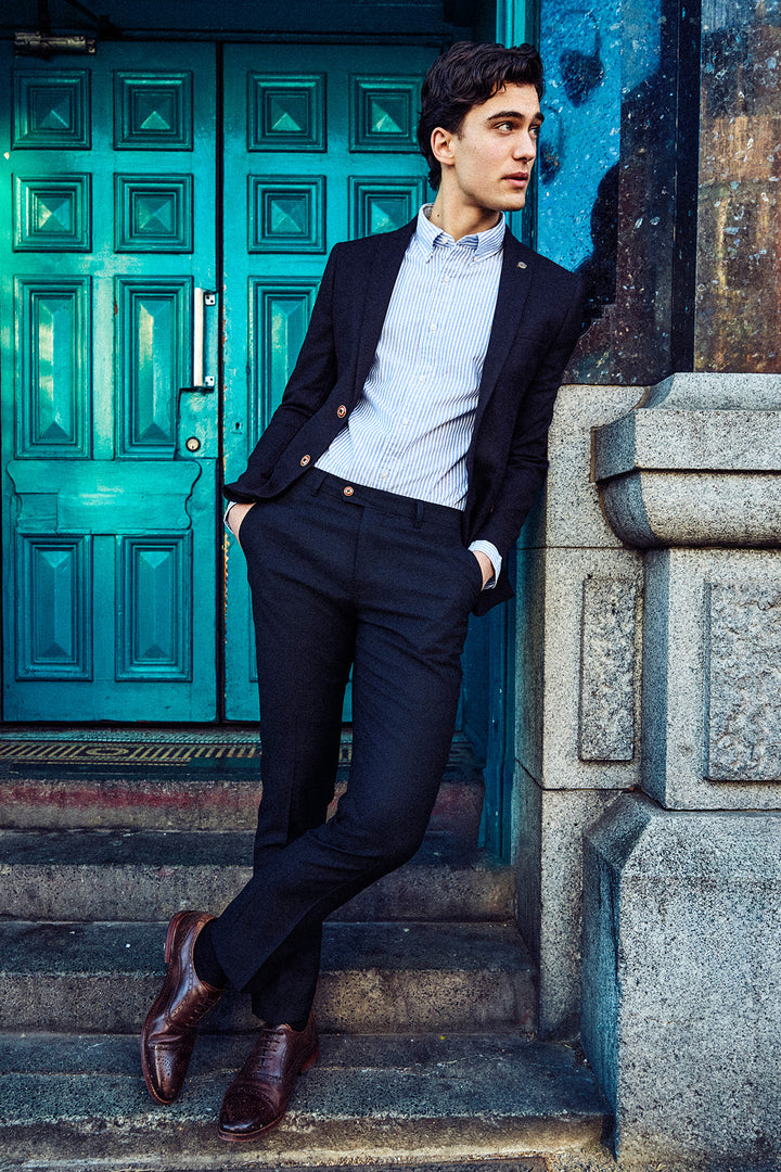 A young man in the Marc Darcy MAX Navy Blue Two Piece Suit leans against a stone pillar by teal double doors.