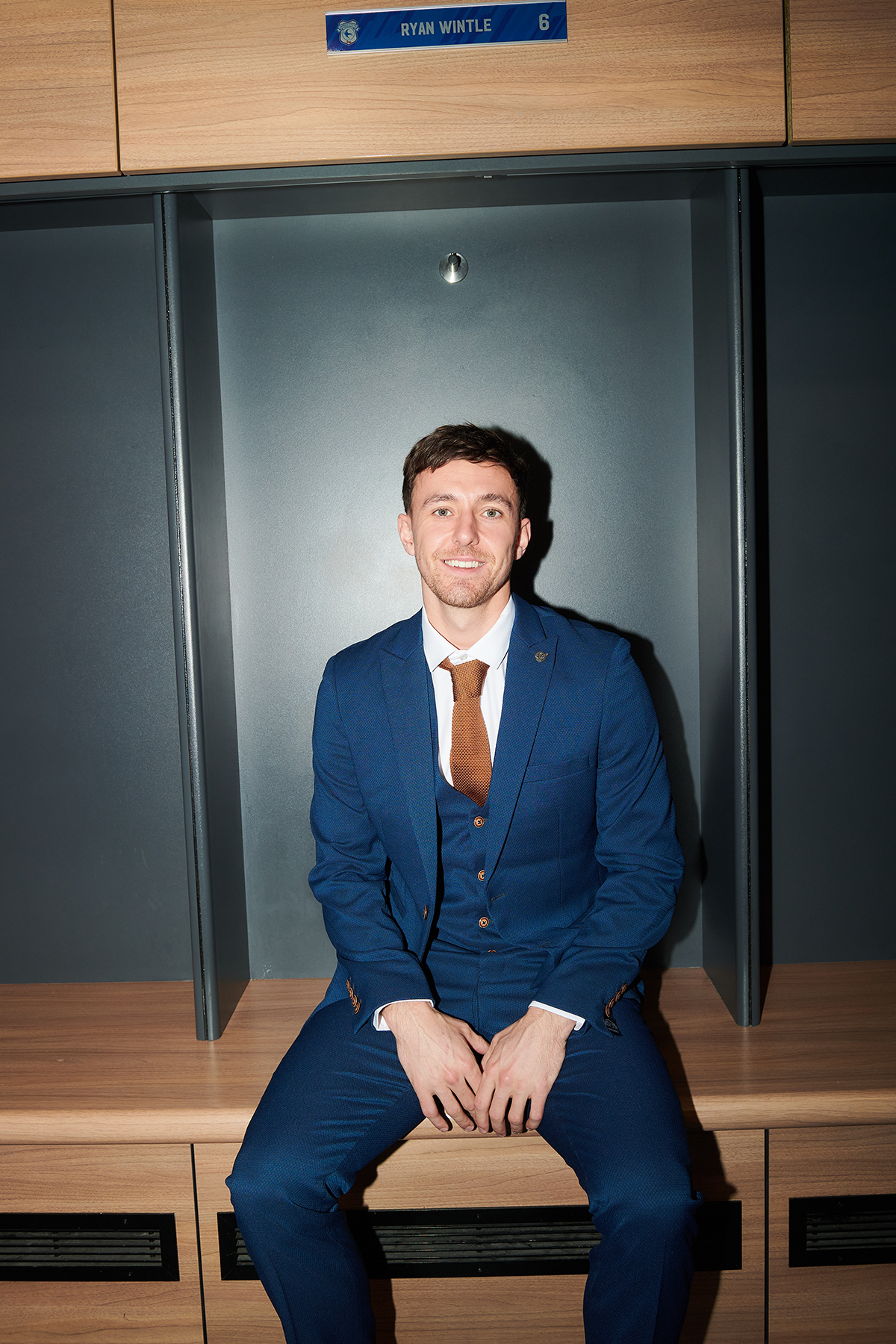 Ryan Wintle smiles at the camera from a locker room bench, dressed in the marcdarcy Official Cardiff City F.C. Club Suit | MAX Royal Blue Suit, paired with a tan tie for a sharp three-piece look.