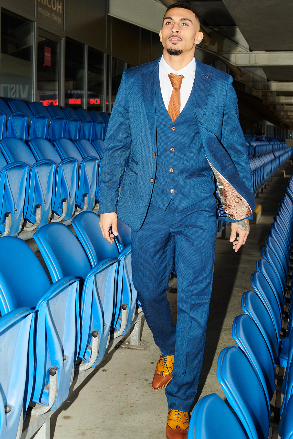 A man in the marcdarcy Official Cardiff City F.C. Club Suit | MAX Royal Blue Suit walks confidently through blue stadium seats, wearing a brown tie and yellow shoes, looking ahead with a slight smile.