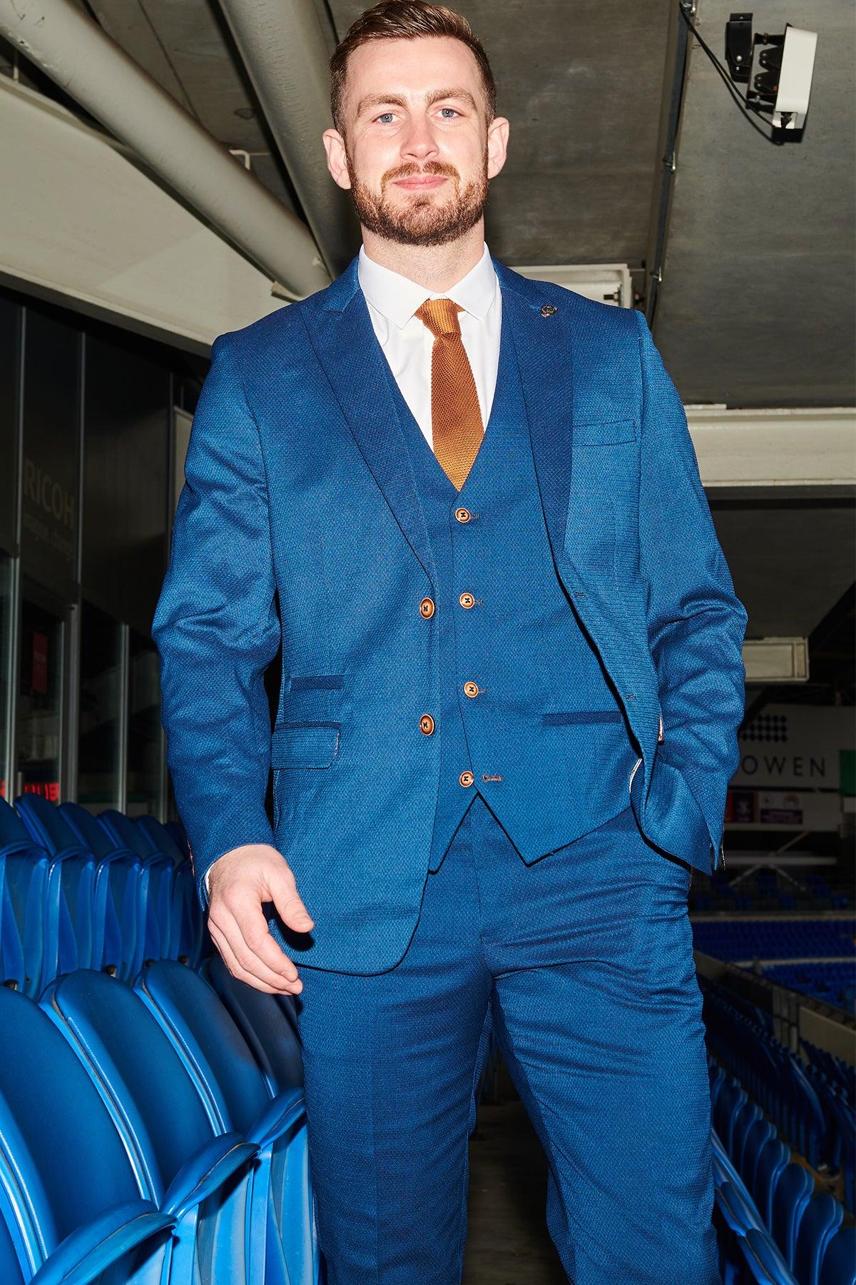 A bearded man smiles at the camera in the stadium seating area, wearing the marcdarcy Official Cardiff City F.C. Club Suit | MAX Royal Blue Suit with a brown tie and white shirt, surrounded by rows of blue seats.