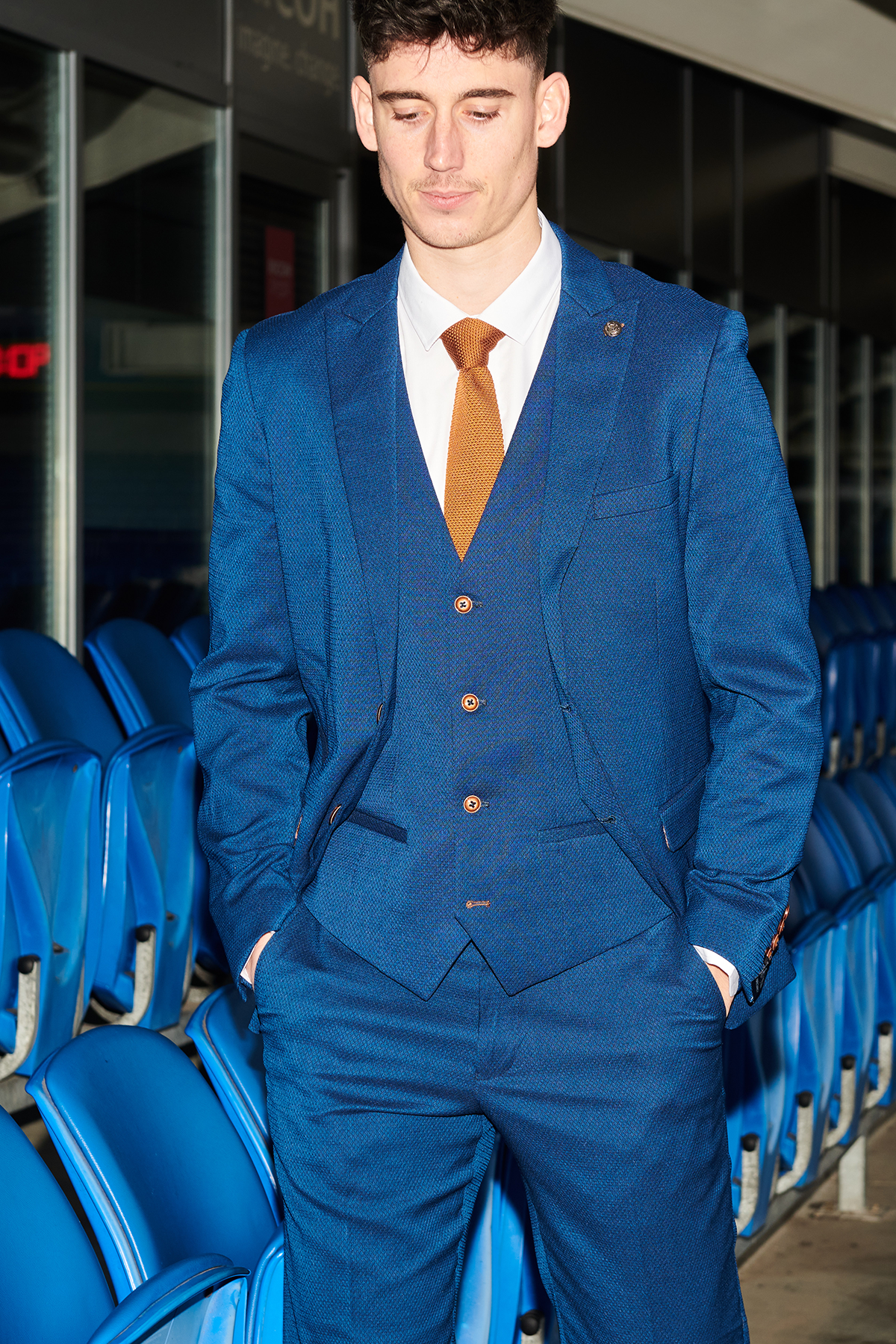 A young man wearing the marcdarcy Official Cardiff City F.C. Club Suit | MAX Royal Blue Suit with a slim fit blazer and orange tie stands with his hands in his pockets before stadium seats.