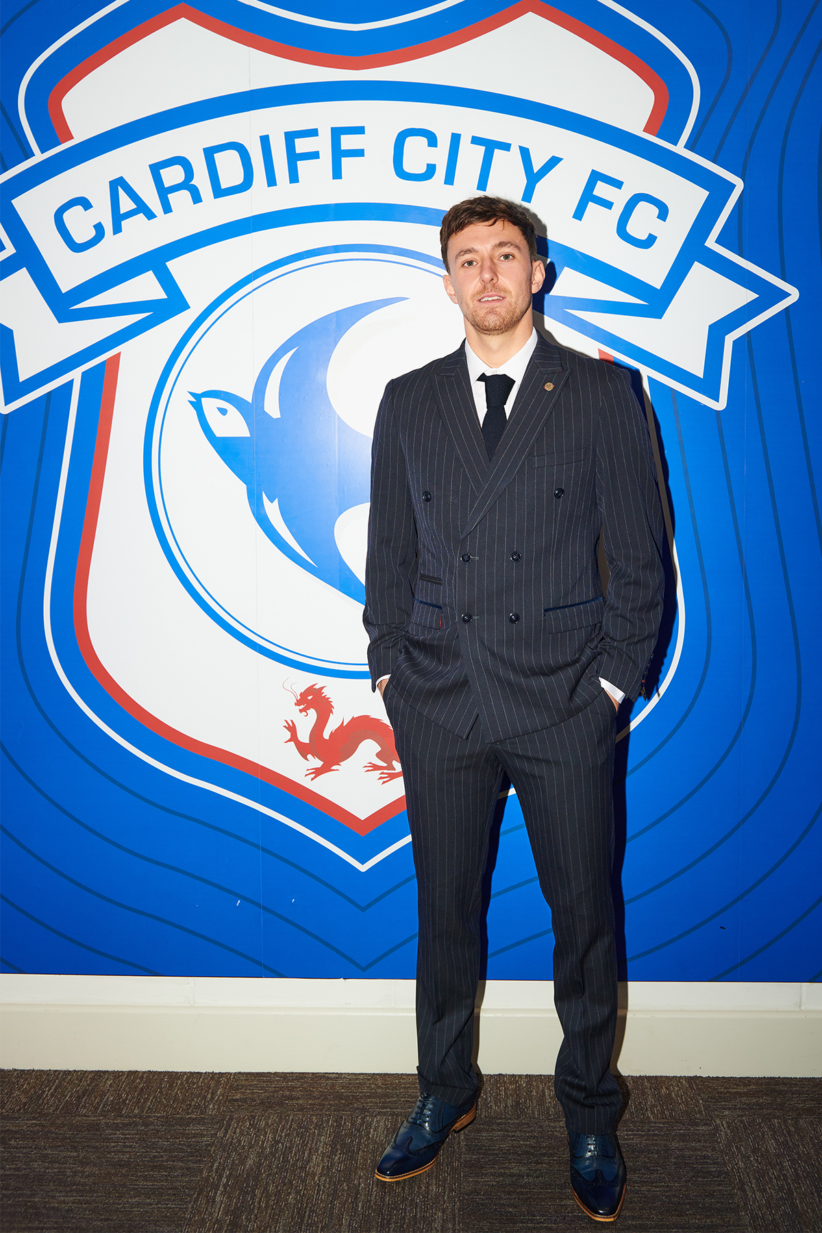 A man wearing the Marc Darcy ROCCO Navy Double Breasted Suit from The Cardiff City F.C. Collection, as worn by Ryan Wintle, stands before a large Cardiff City FC logo on a blue wall, hands in pockets and dressed with a tie.