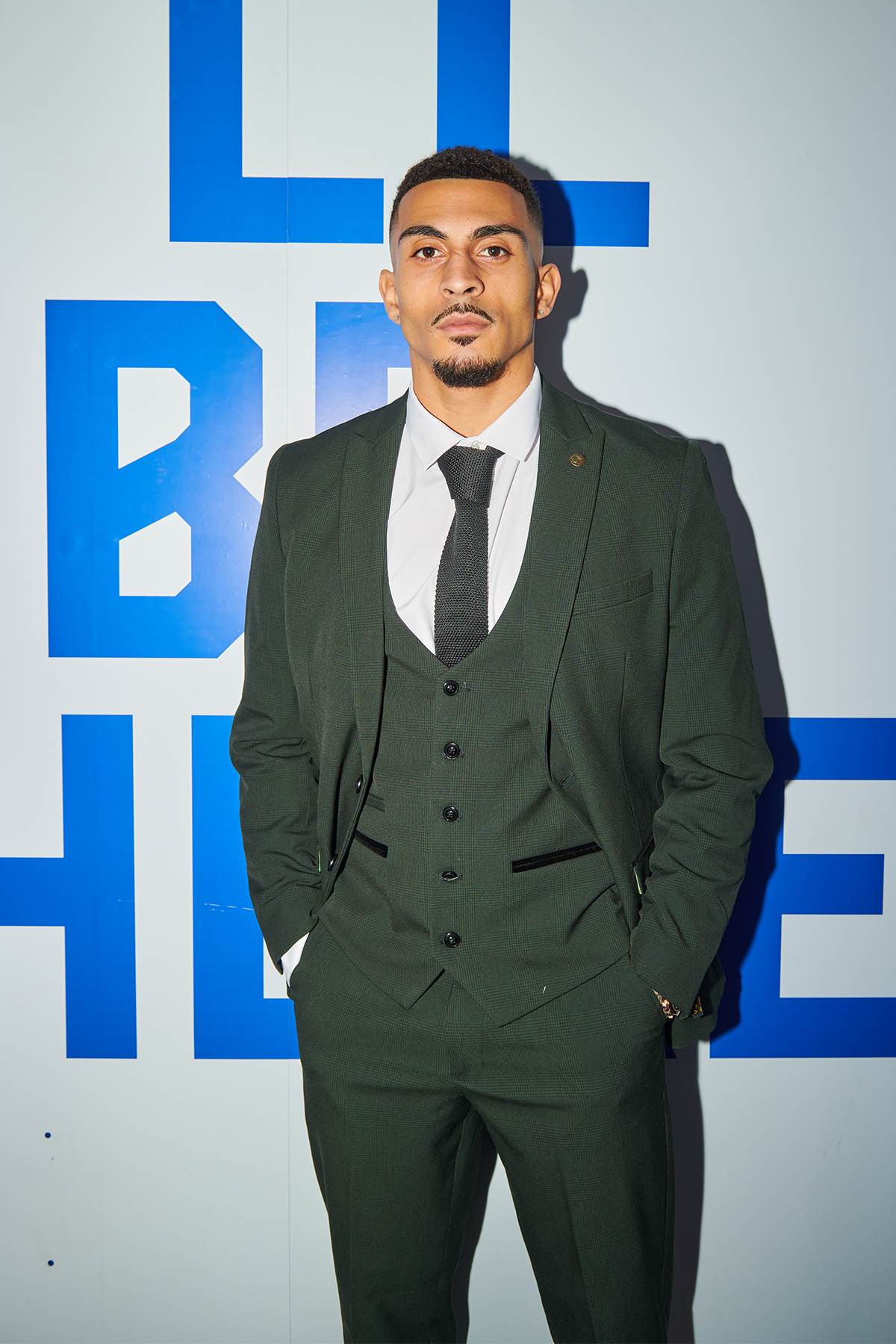 A man wears the Marc Darcy BROMLEY Olive Green Check Suit from The Cardiff City F.C. Collection, as worn by Karlan Grant. He stands before a white wall with large blue letters, sporting short hair, a trimmed beard, and hands in his pockets.