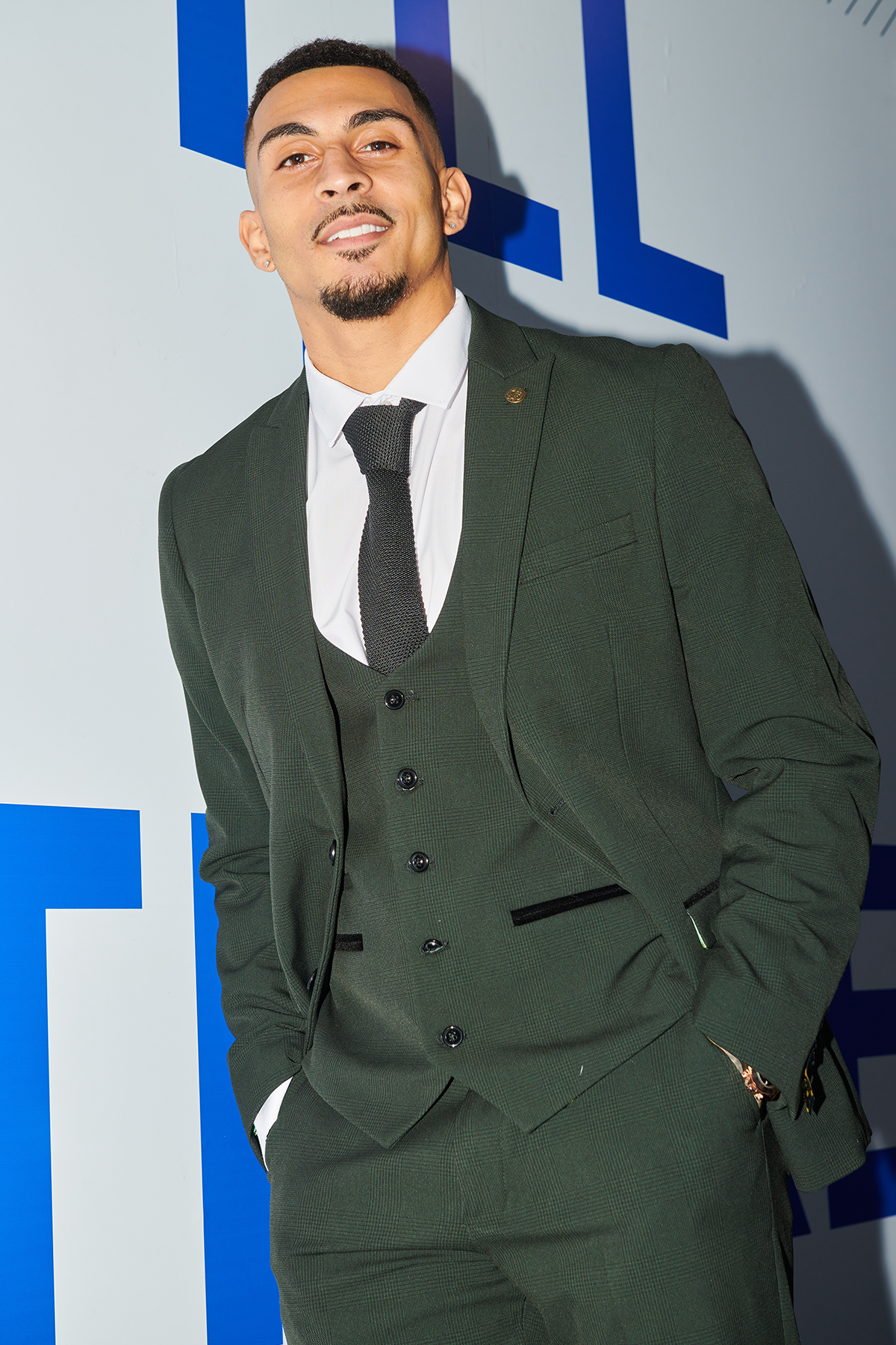 A young man smiles at an event, hands in pockets, wearing the Marc Darcy Cardiff City F.C. Collection - BROMLEY Olive Green Check Suit as worn by Karlan Grant, paired with a white shirt and black knit tie, in front of a blue and white backdrop.
