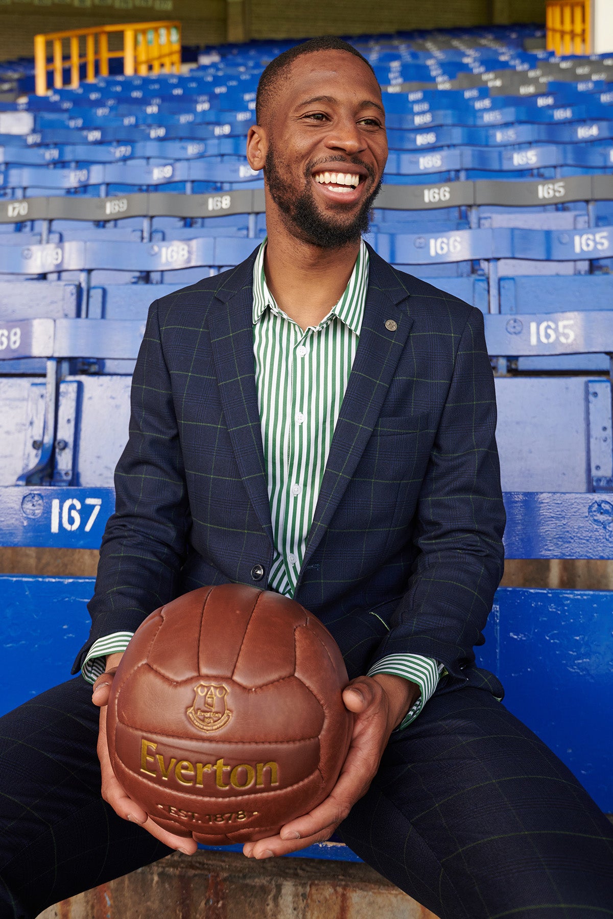 A man wears the Marc Darcy The Everton Collection Beto In EDINSON Navy Green Check Two Piece Suit, paired with a striped shirt, while seated in stadium seats holding an Everton football.