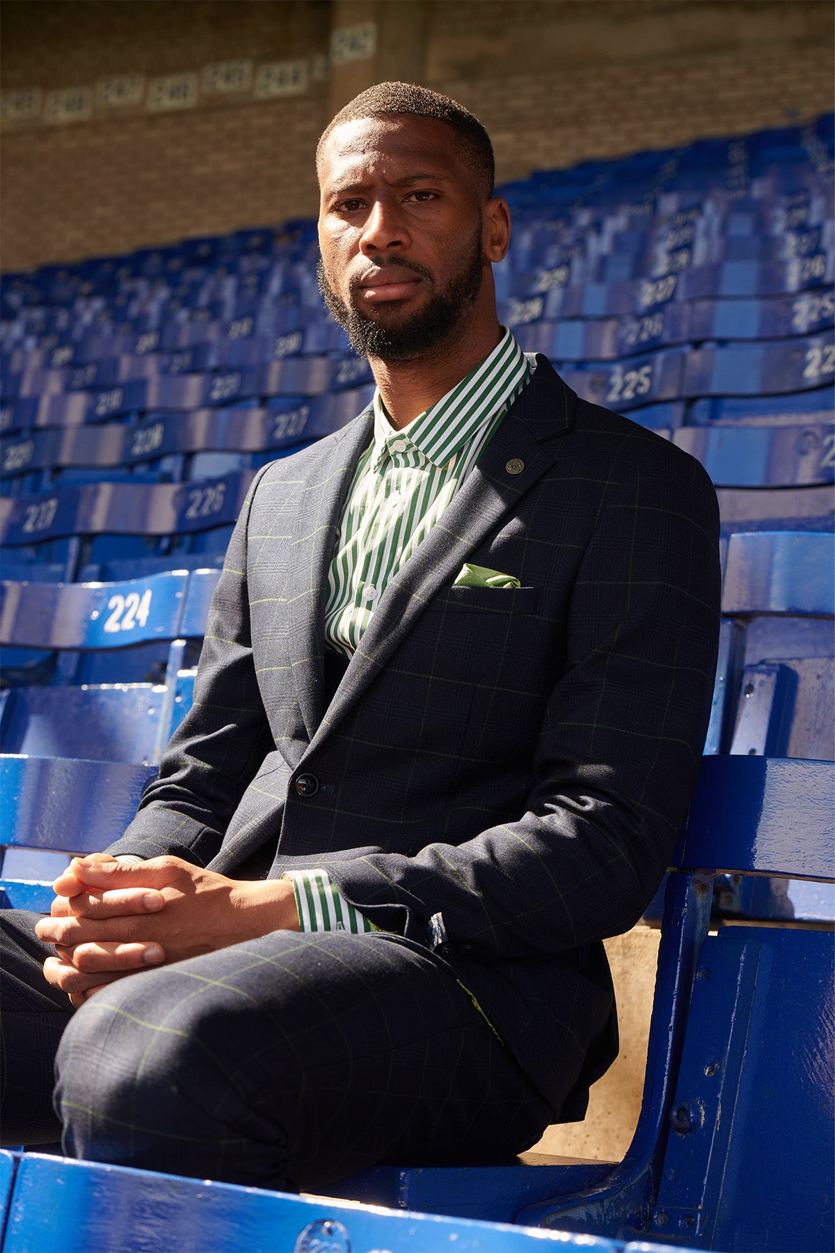 A man in the Marc Darcy The Everton Collection | Beto In EDINSON Navy Green Check Two Piece Suit sits on blue stadium seats, wearing a green and white striped shirt. Sunlight highlights his face and hands amid empty surrounding seats as he looks at the camera.