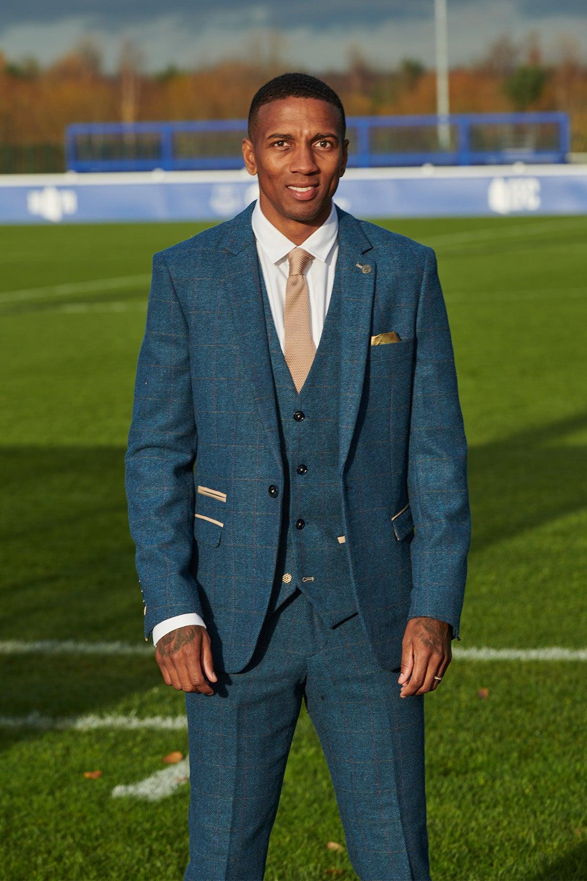 Ashley Young wears the marcdarcy Everton Collection DION Blue Tweed Check Suit—a slim-fit three-piece—paired with a white shirt and beige tie, standing on a grassy sports field with white markings and a blue fence in the background.