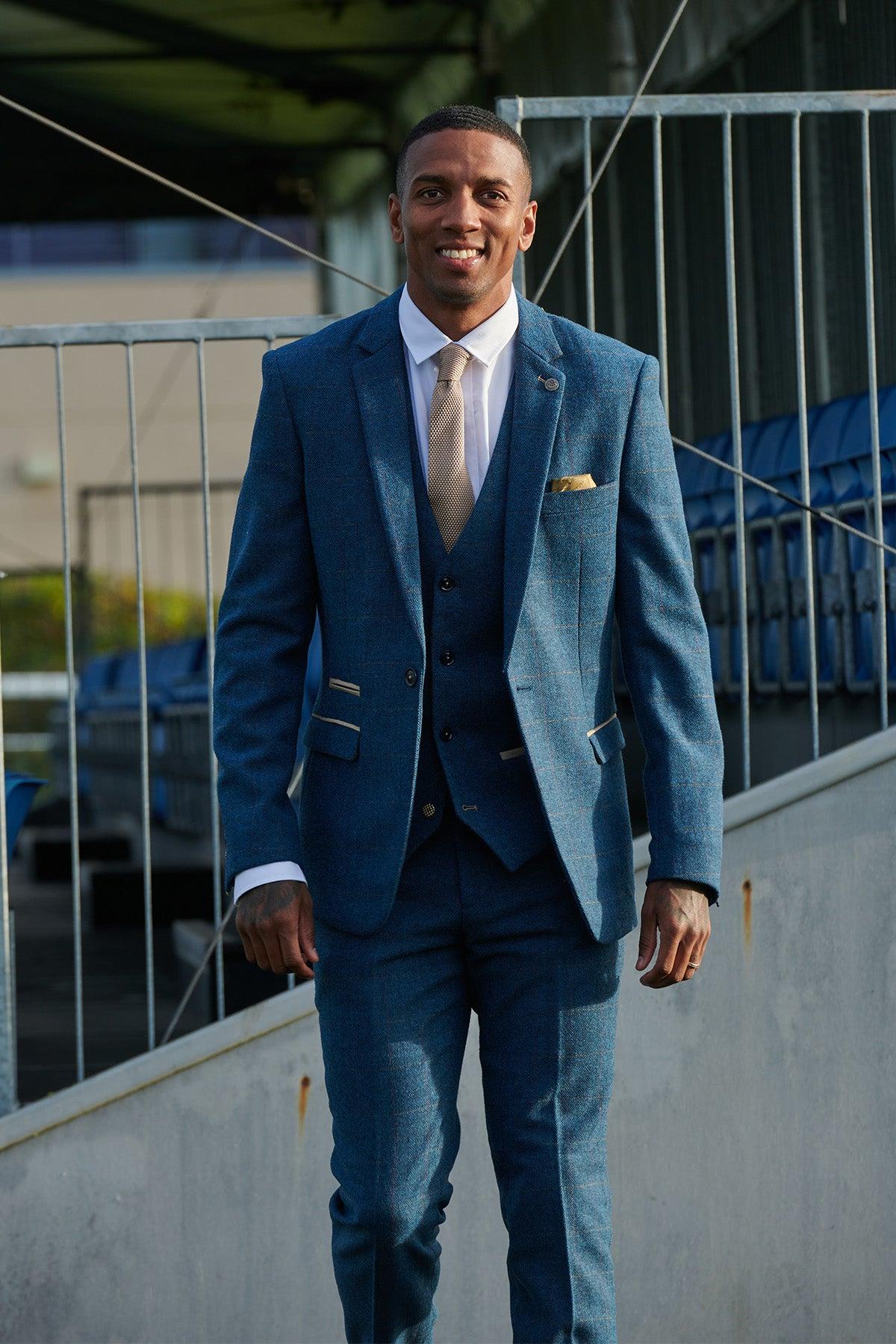 Ashley Young smiles at the camera in the marcdarcy Everton Collection DION Blue Tweed Check Suit, paired with a white shirt and tan tie, standing before stadium seating.