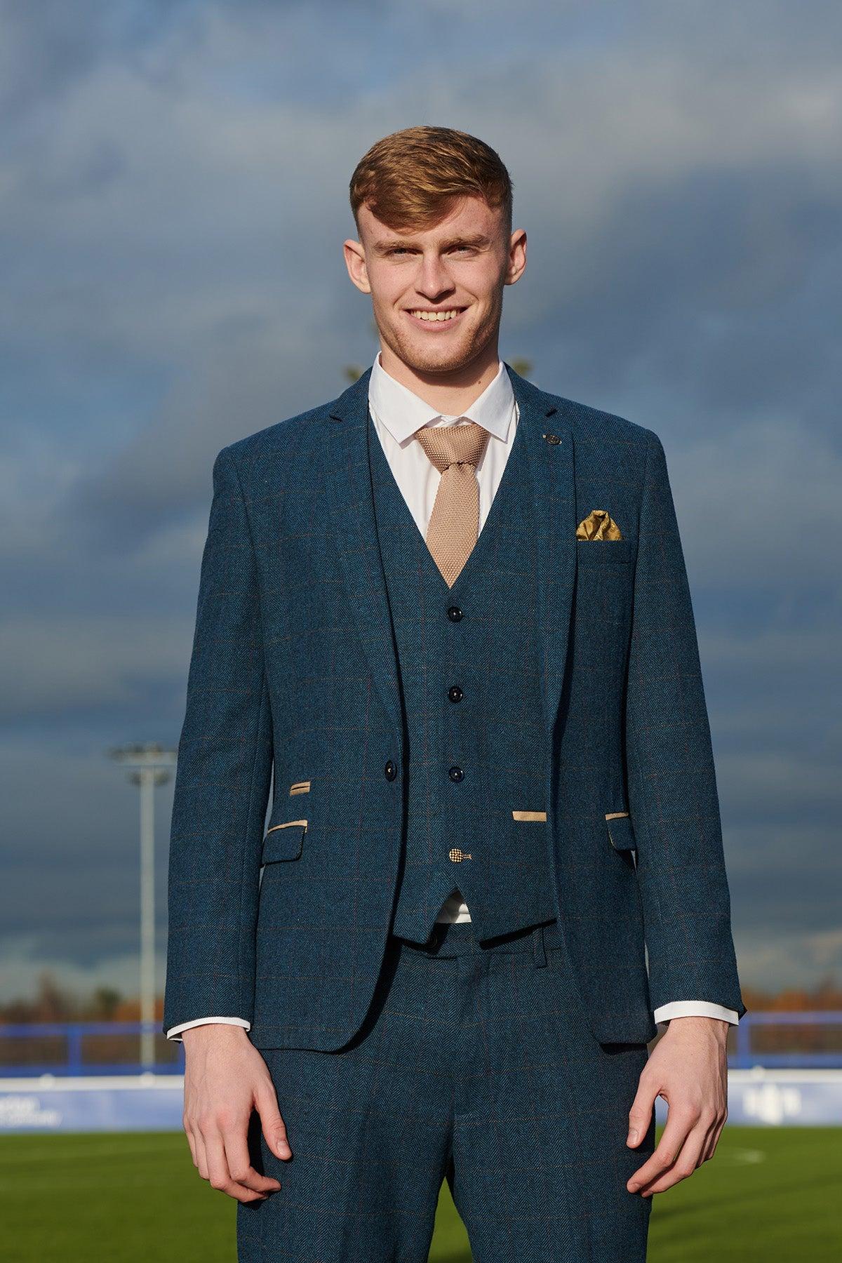 Jarrad Branthwaite, outdoors on a cloudy day by a sports field, smiles in The Everton Collection | DION Blue Tweed Check Suit by marcdarcy, styled with a tan tie and pocket square.