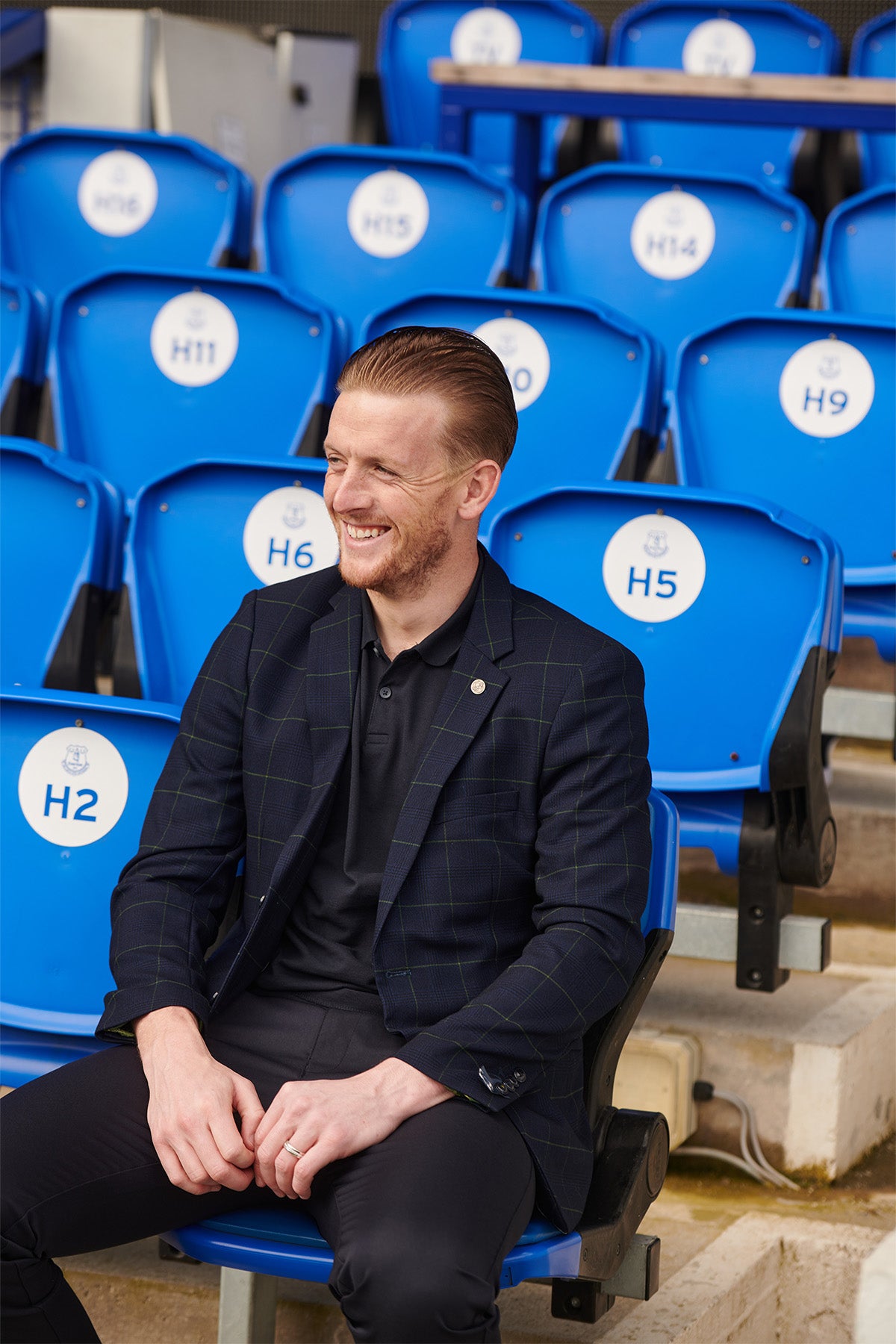 A man wearing the Marc Darcy Everton Collection's Edinson Navy Green Check Blazer smiles while sitting in blue stadium seats.