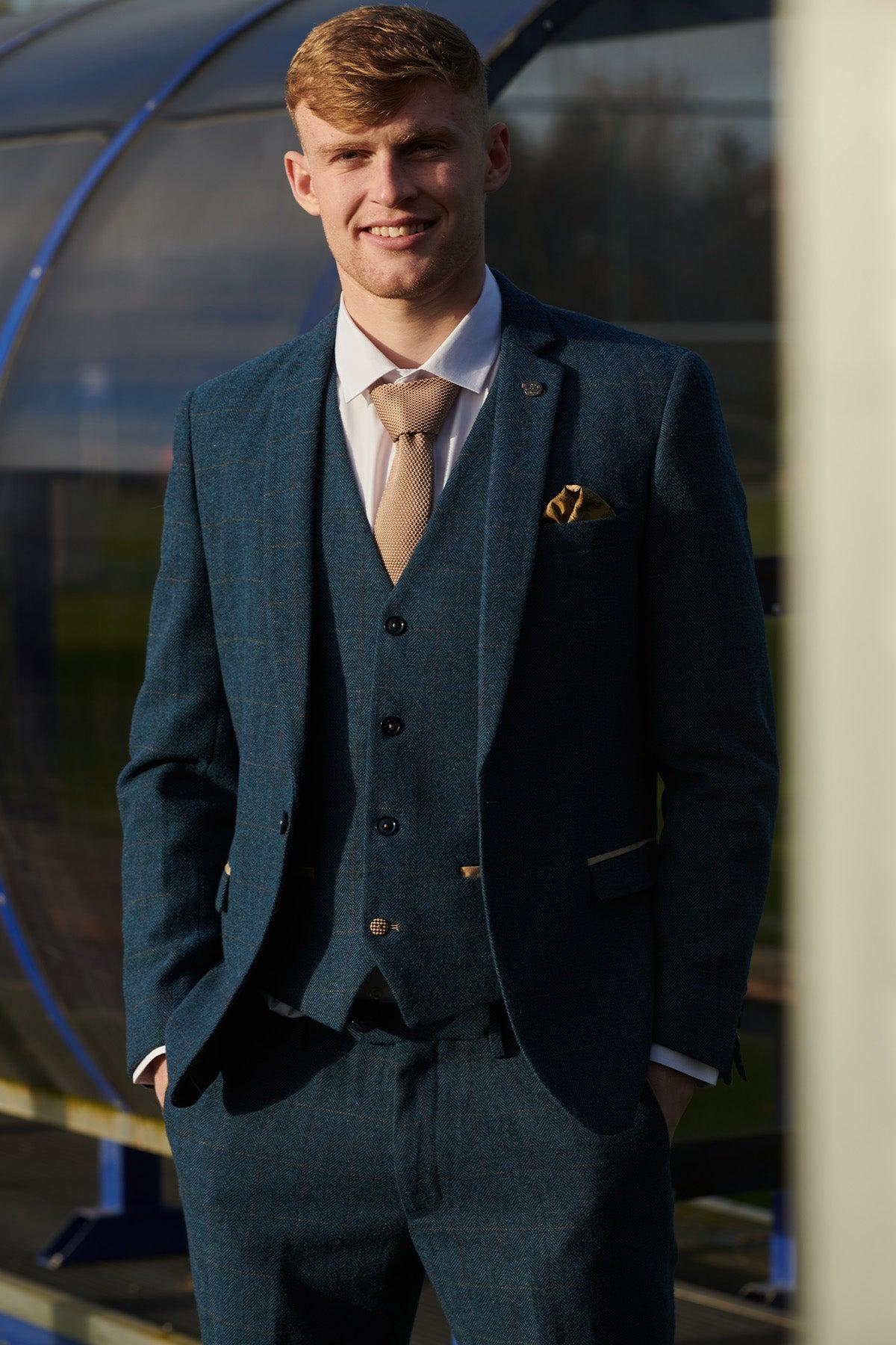 A man wears the marcdarcy Everton Collection Jarrad Branthwaite DION Blue Tweed Check Suit with a light shirt and beige tie, smiling outdoors with a blurred structure and sunlight in the background.