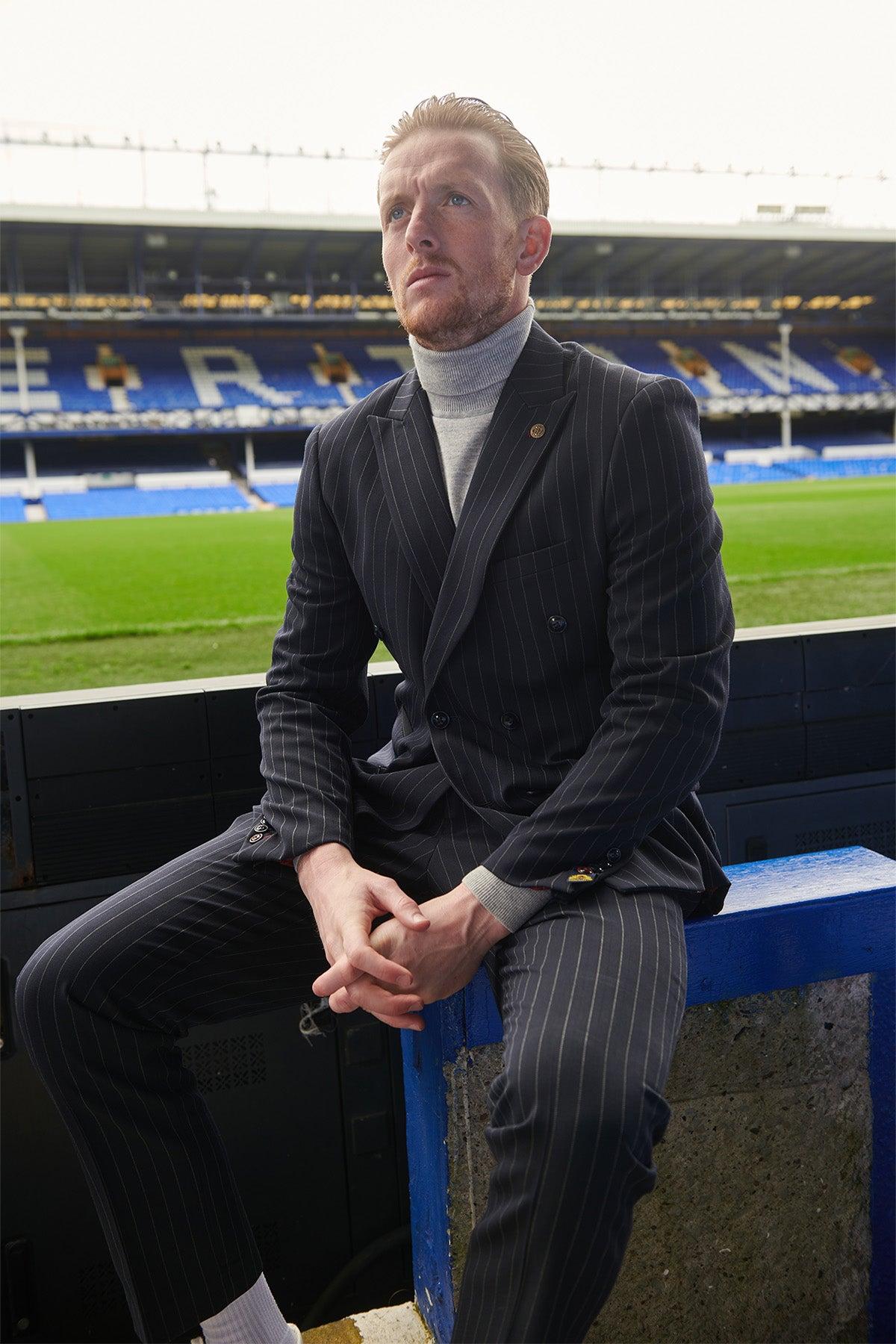 A man wearing The Everton Collection | Jordan Pickford in ROCCO Navy Pinstripe Double Breasted Two Piece Suit by Marc Darcy and a turtleneck sits on a blue bench at a soccer stadium.