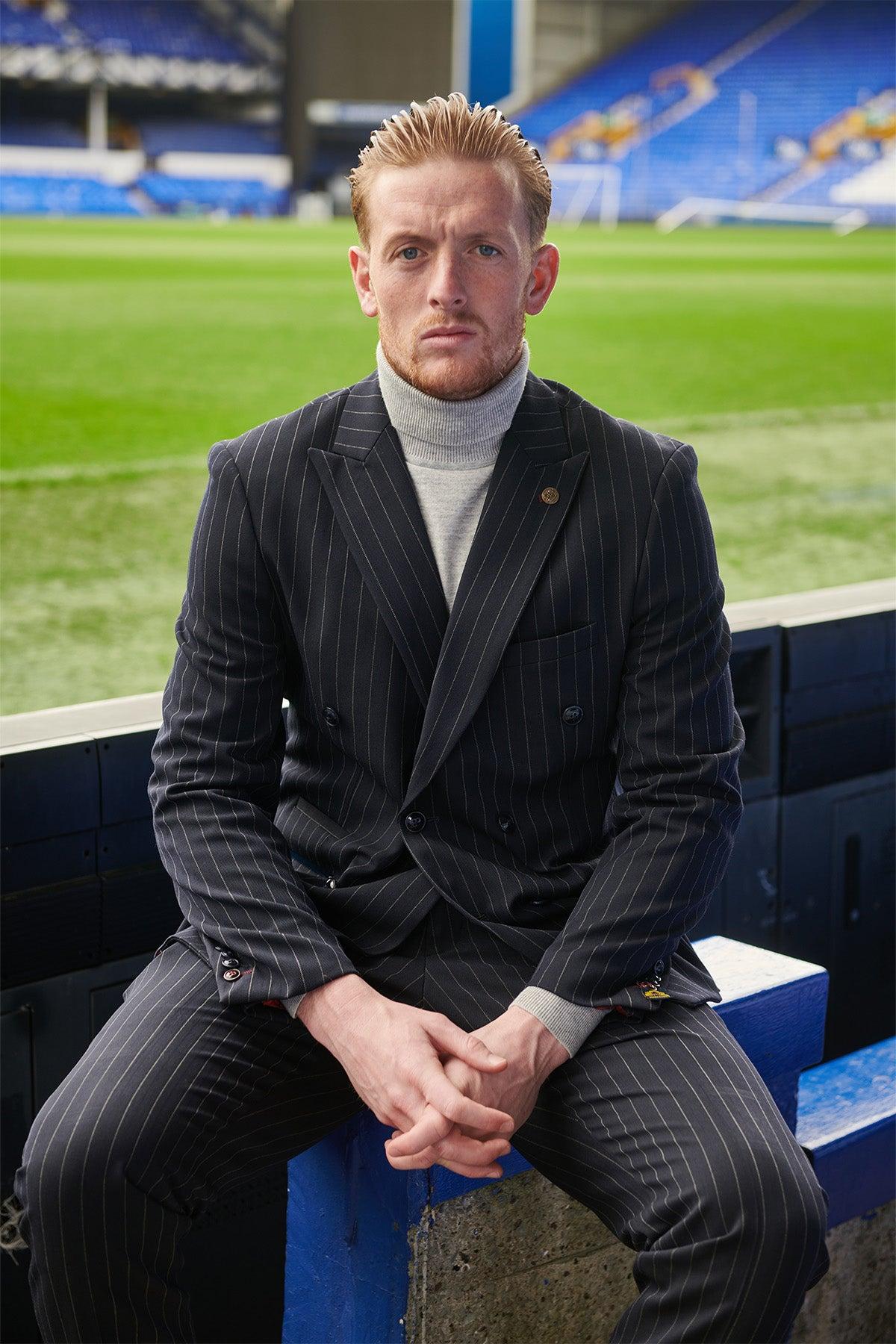 A man wearing the Marc Darcy ROCCO Navy Pinstripe Double Breasted Suit from The Everton Collection sits in a stadium with his hands clasped, inspired by Jordan Pickford.
