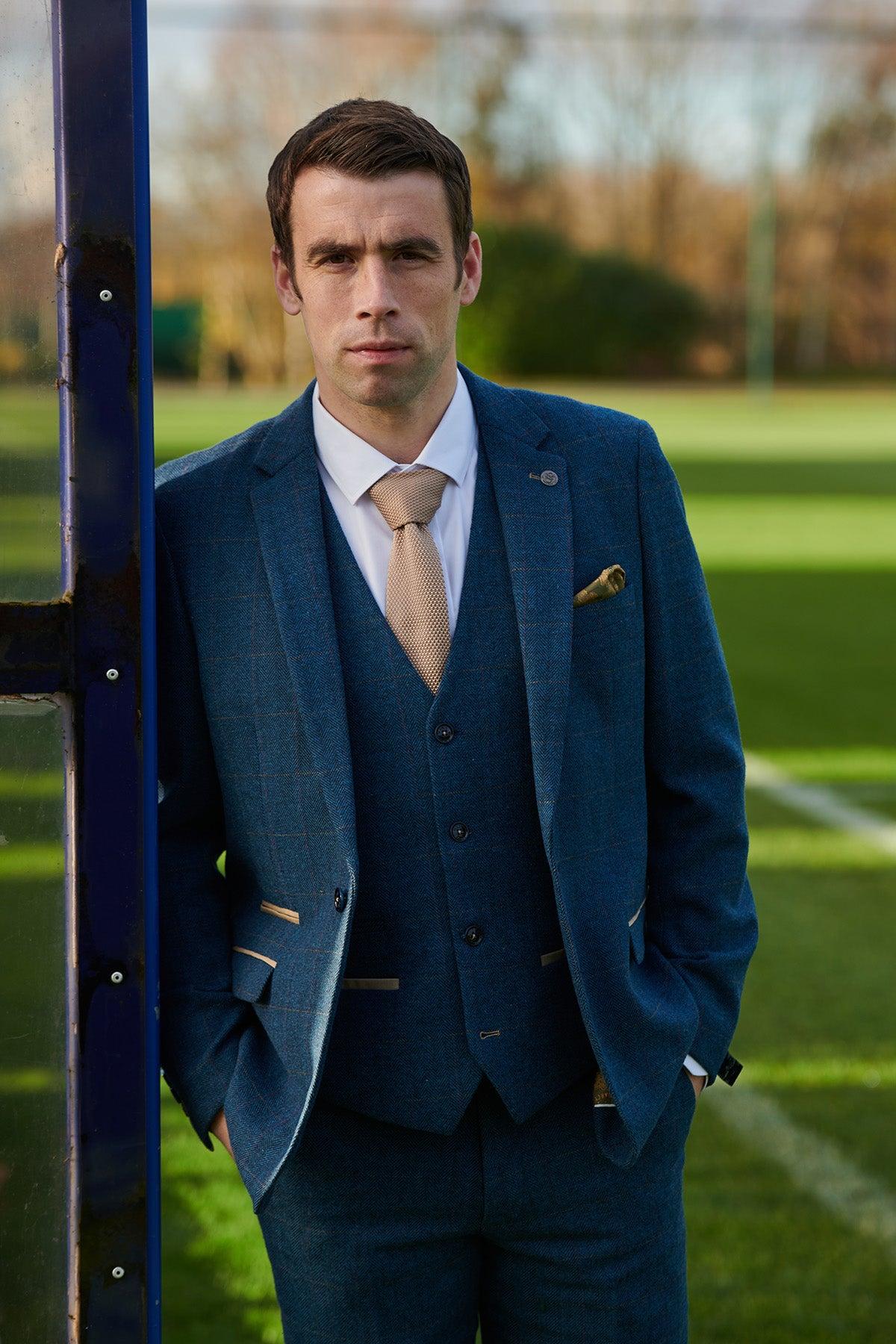 A man in marcdarcy’s Séamus Coleman DION Blue Tweed Check Suit (The Everton Collection) stands by a blue post on a green field with white markings, wearing a white shirt and beige tie.