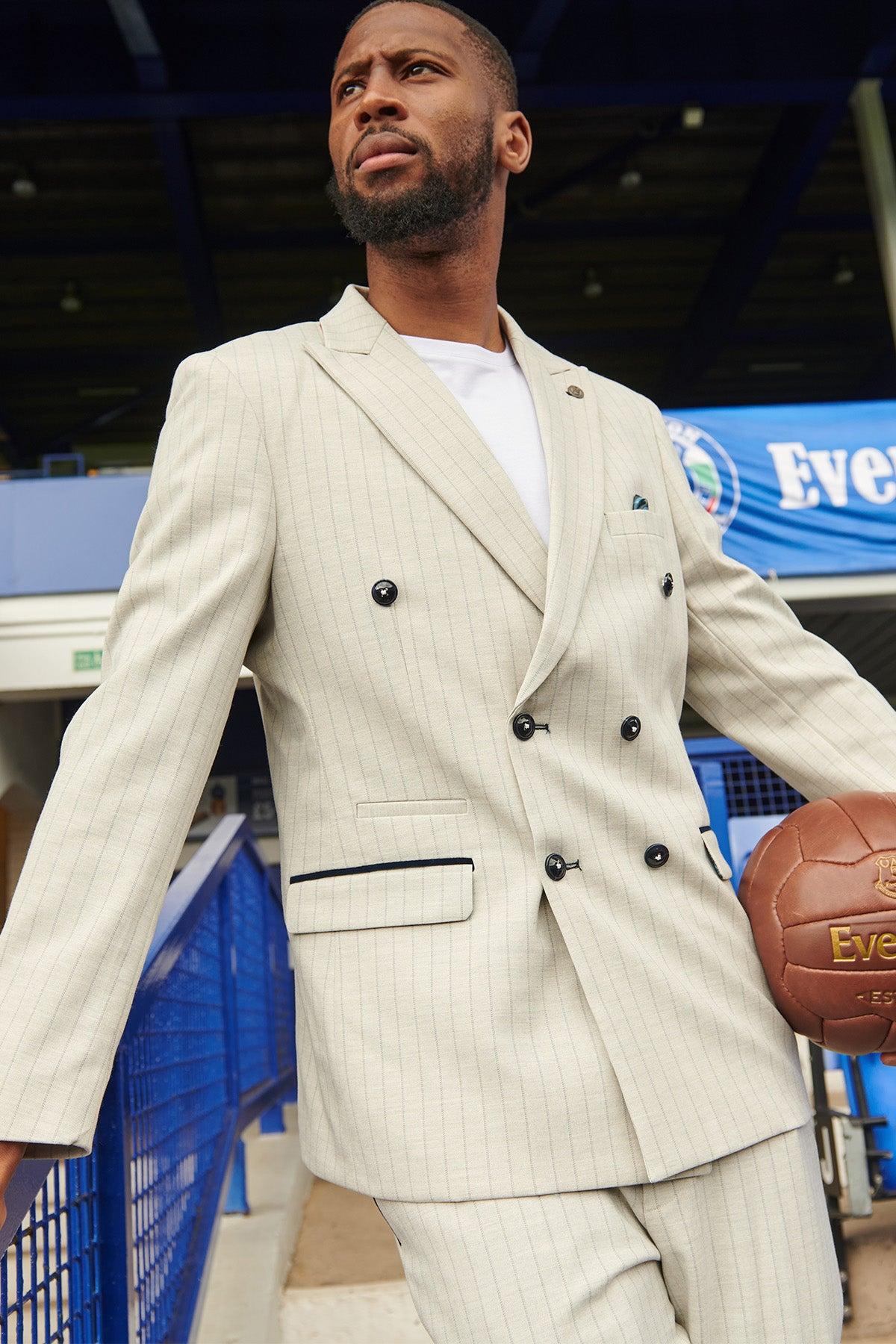 A man in The Everton Collection | Beto In GRANT Stone Pinstripe Double Breasted Suit by Marc Darcy holds a vintage brown soccer ball at a stadium, blue railings and an Everton banner visible in the background.