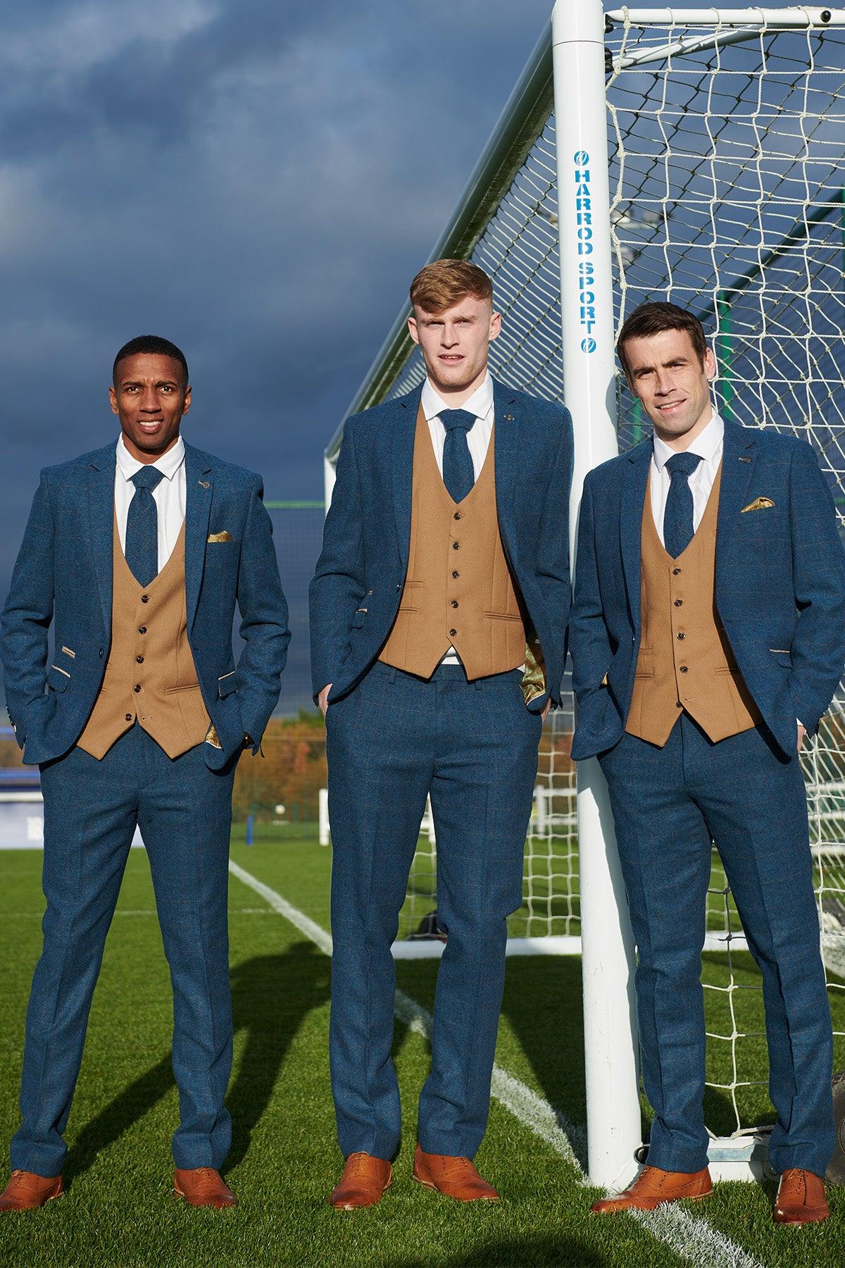 Three men stand by a soccer goal, wearing marcdarcy's The Everton Collection | DION Blue Tweed Suit with Kelvin Waistcoat Team Suit—an ensemble inspired by classic Everton F.C. style, featured under cloudy skies and sunlight.