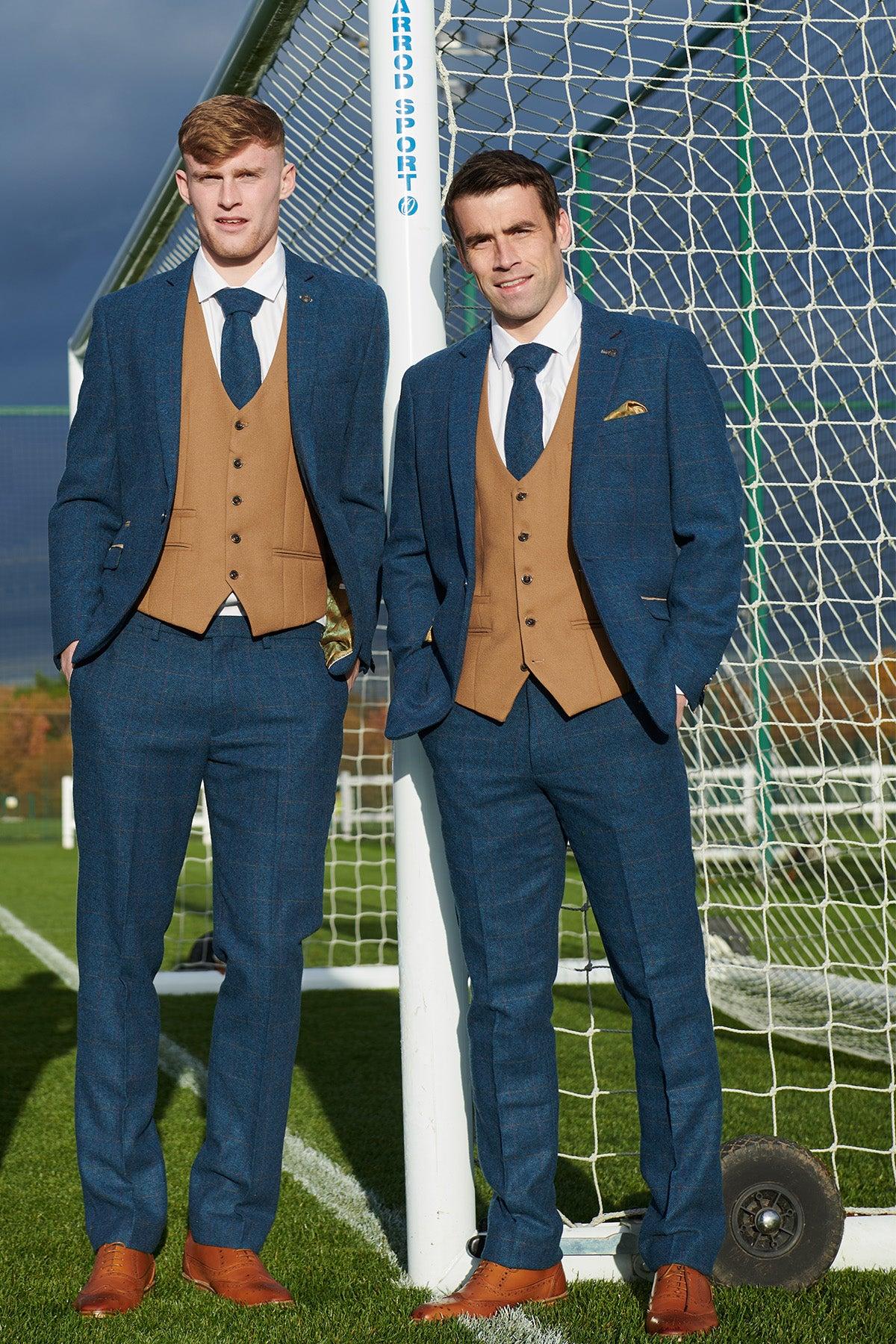 Two men in marcdarcy's The Everton Collection | DION Blue Tweed Suit with Kelvin Waistcoat team suit, white shirts, navy ties, and brown shoes stand on a grass field in front of a soccer goal, posing for a photo.