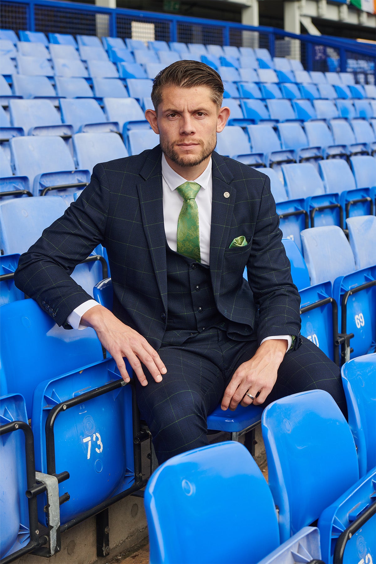 James Tarkowski wears Marc Darcy’s EDINSON Navy Green Check Suit from The Everton Collection, sitting on blue stadium seats with a green tie and pocket square, looking seriously at the camera as seat numbers are visible.