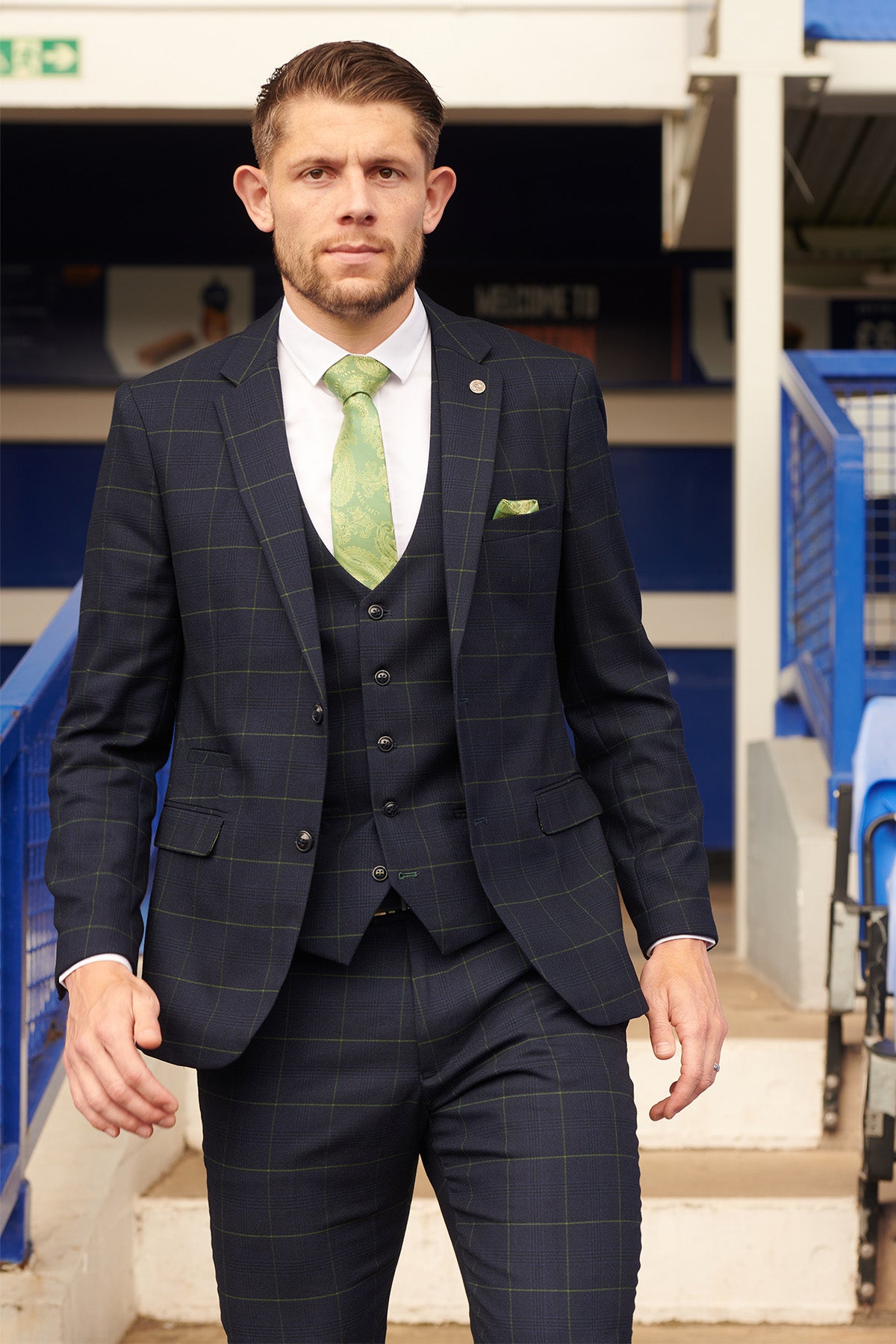 James Tarkowski wears the Marc Darcy EDINSON Navy Green Check Suit from The Everton Collection with a white shirt and green patterned tie, standing outdoors before blue stadium seats and railings.