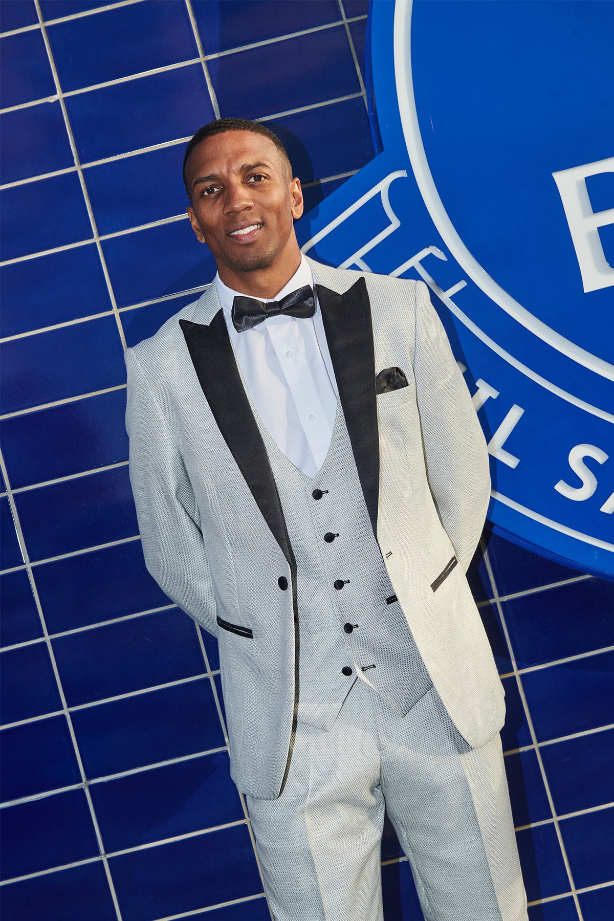Ashley Young wears the marcdarcy SPENCER Stone Tux Lapel Suit from The Everton Collection, paired with a black bow tie, smiling with hands behind his back in front of a blue tiled wall and large circular sign.