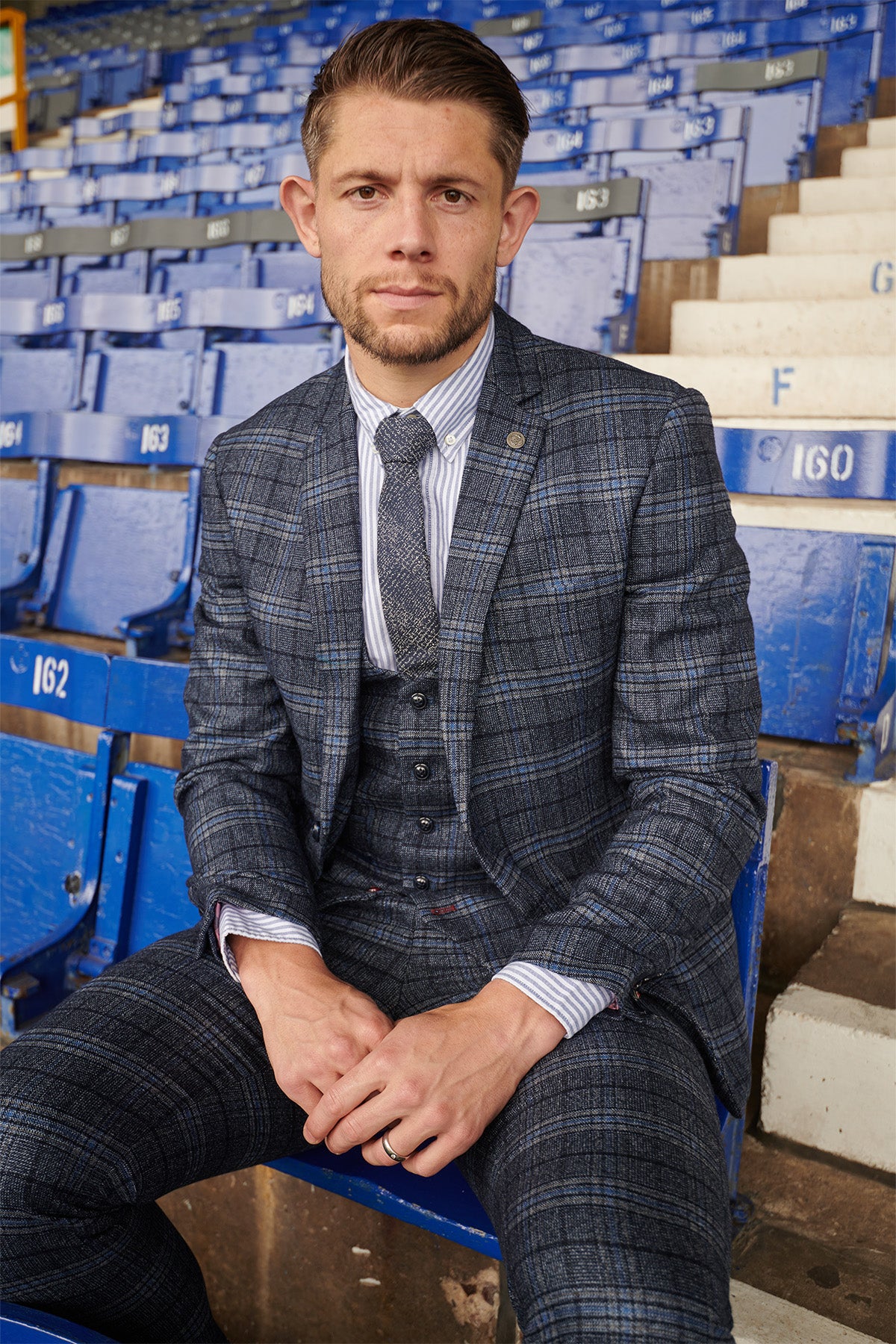 James Tarkowski wears the Marc Darcy ANDY Blue Check Suit from The Everton Collection while sitting on blue stadium seats.