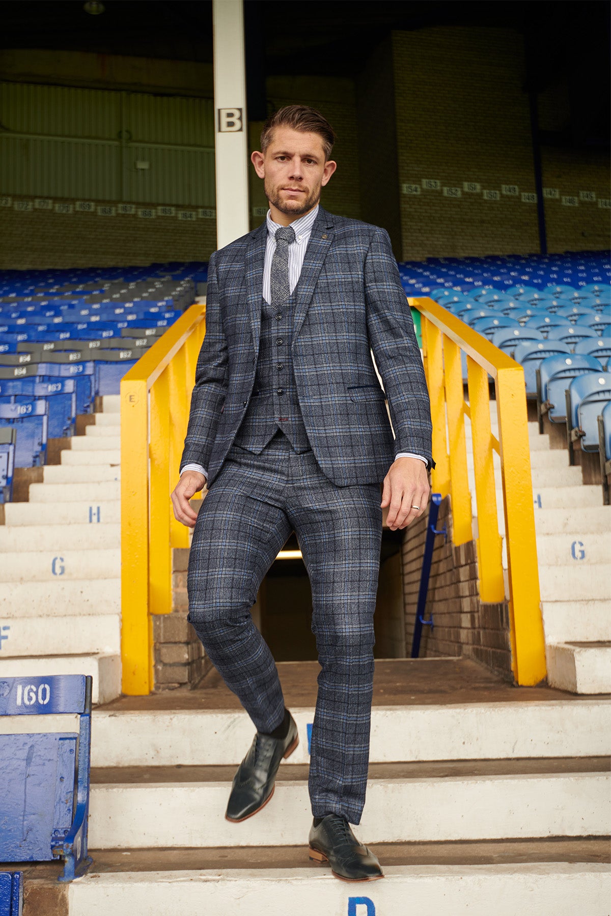 James Tarkowski, wearing the ANDY Blue Check Suit from Marc Darcy's The Everton Collection, walks down stadium steps with yellow railings and empty blue seats in the background.