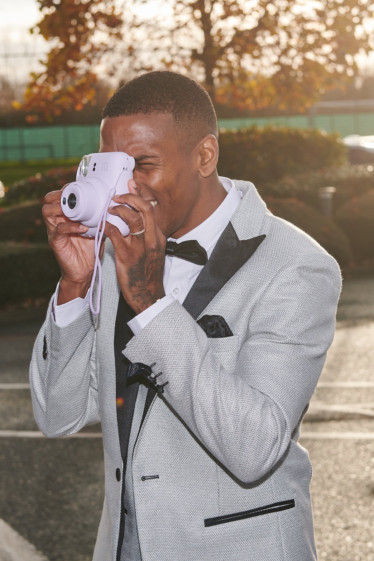 A man wearing marcdarcy’s The Everton Collection | Ashley Young In SPENCER Stone Tux Lapel Suit smiles as he takes a photo with a lilac instant camera, paired with a black bow tie.