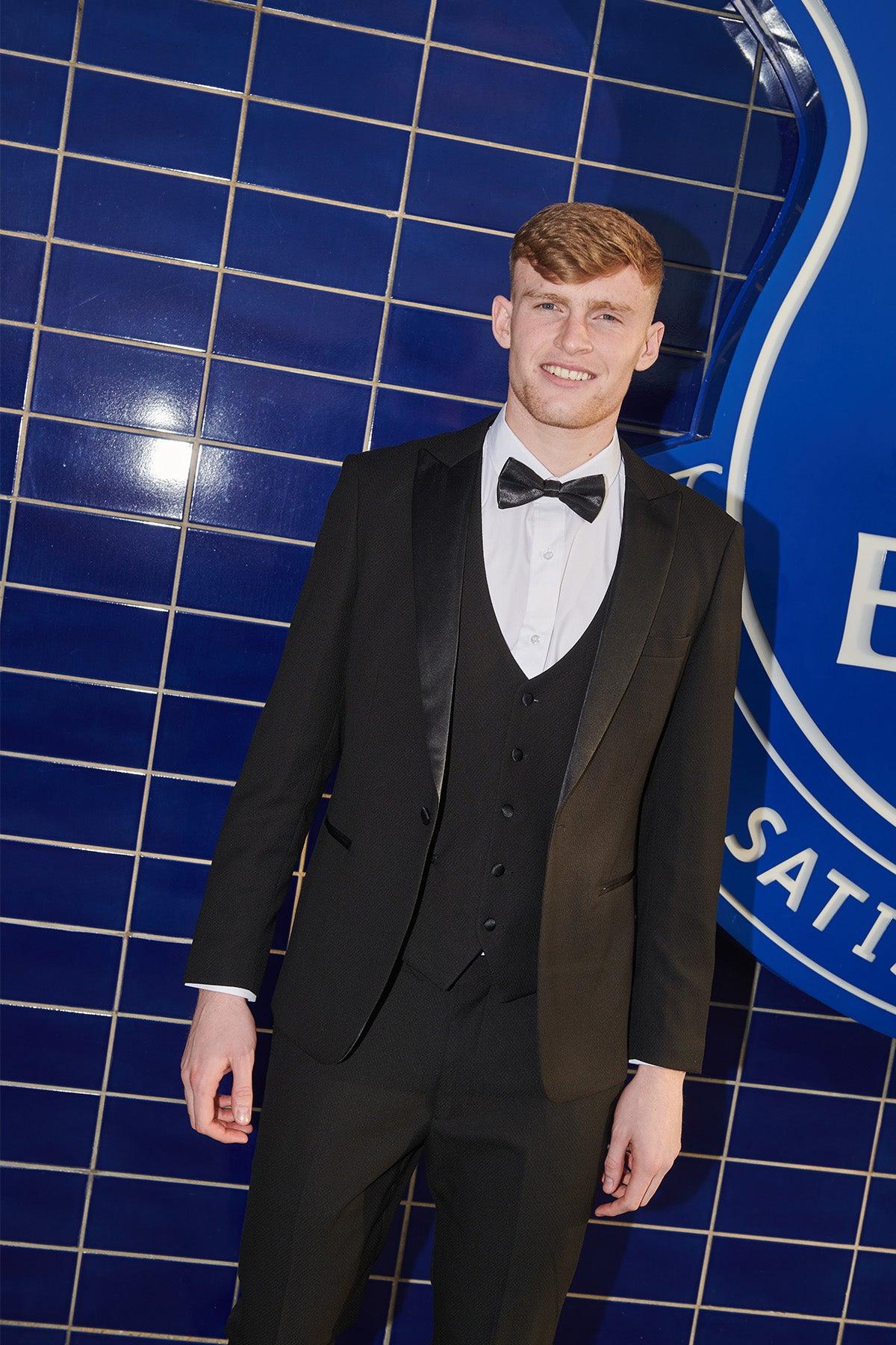 Jarrad Branthwaite wears the marcdarcy DALTON Black Tux Lapel Diamond Suit from The Everton Collection, smiling in front of a dark blue tiled wall with a partial blue and white emblem behind him.