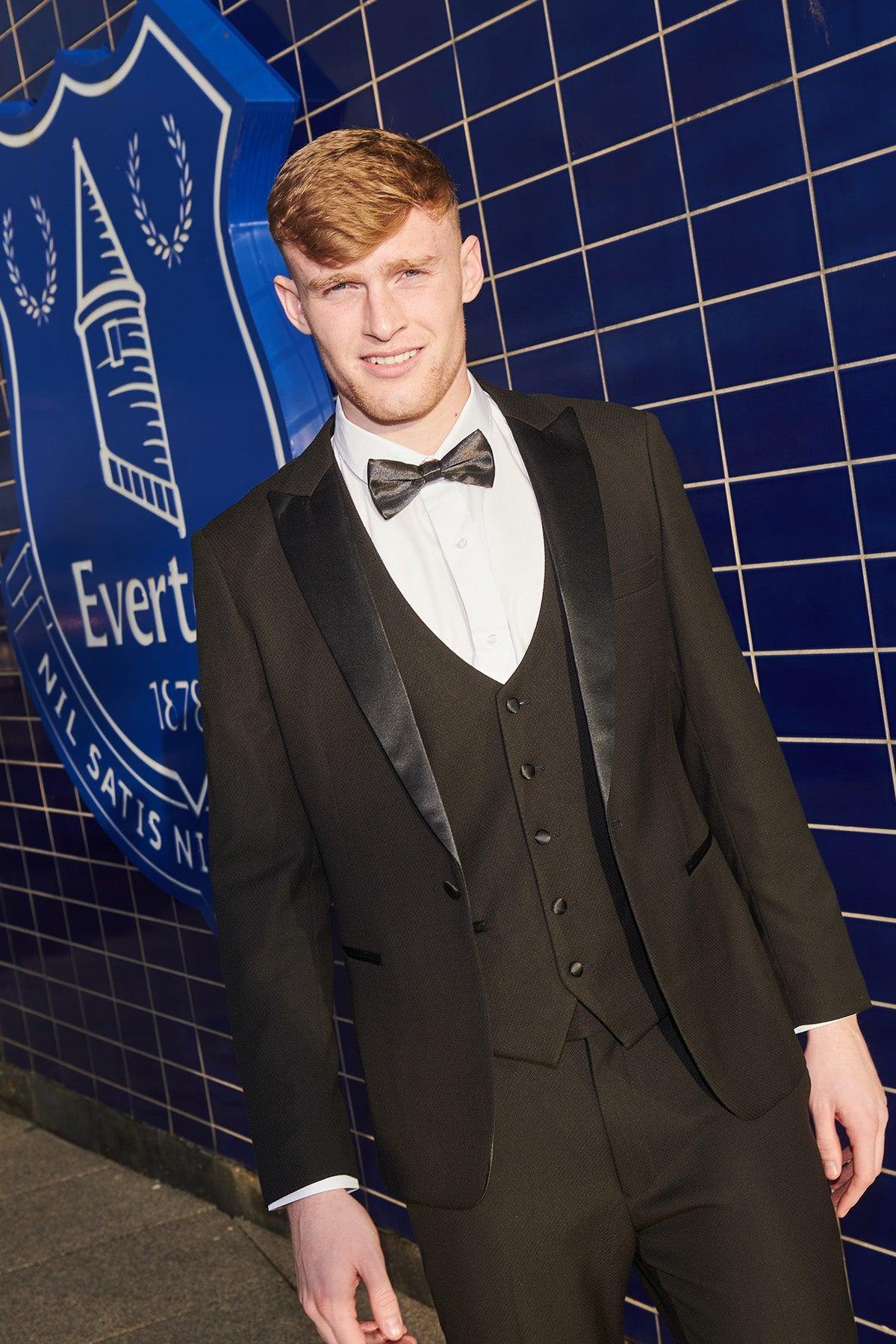 Jarrad Branthwaite wears the marcdarcy DALTON Black Tux Lapel Diamond Suit from The Everton Collection, smiling in front of a blue tiled wall with the Everton Football Club crest.