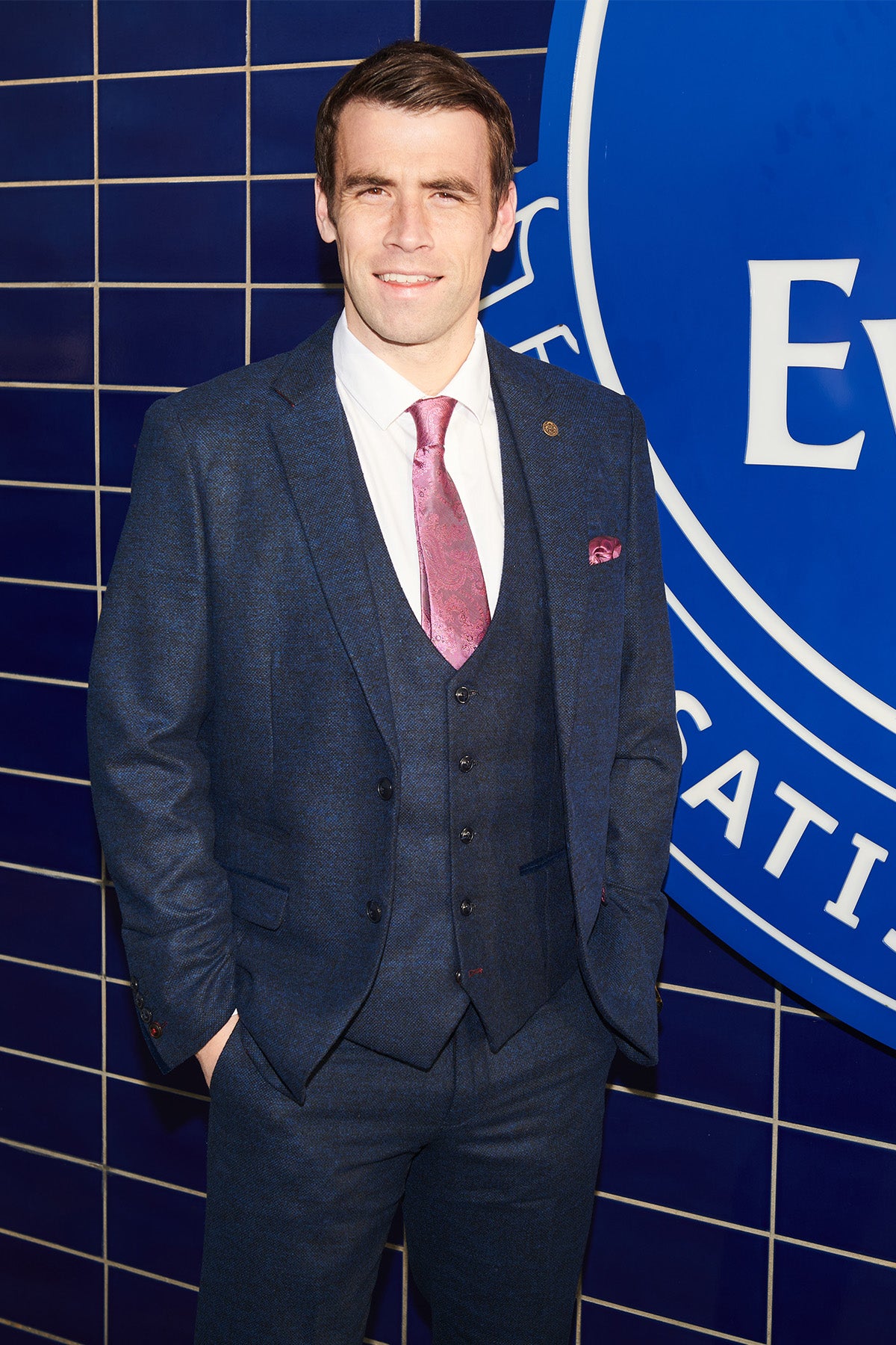 Séamus Coleman smiles at the camera wearing the marcdarcy MARLOW Navy Tweed Suit from The Everton Collection, hands in his pockets, paired with a white shirt and pink patterned tie.
