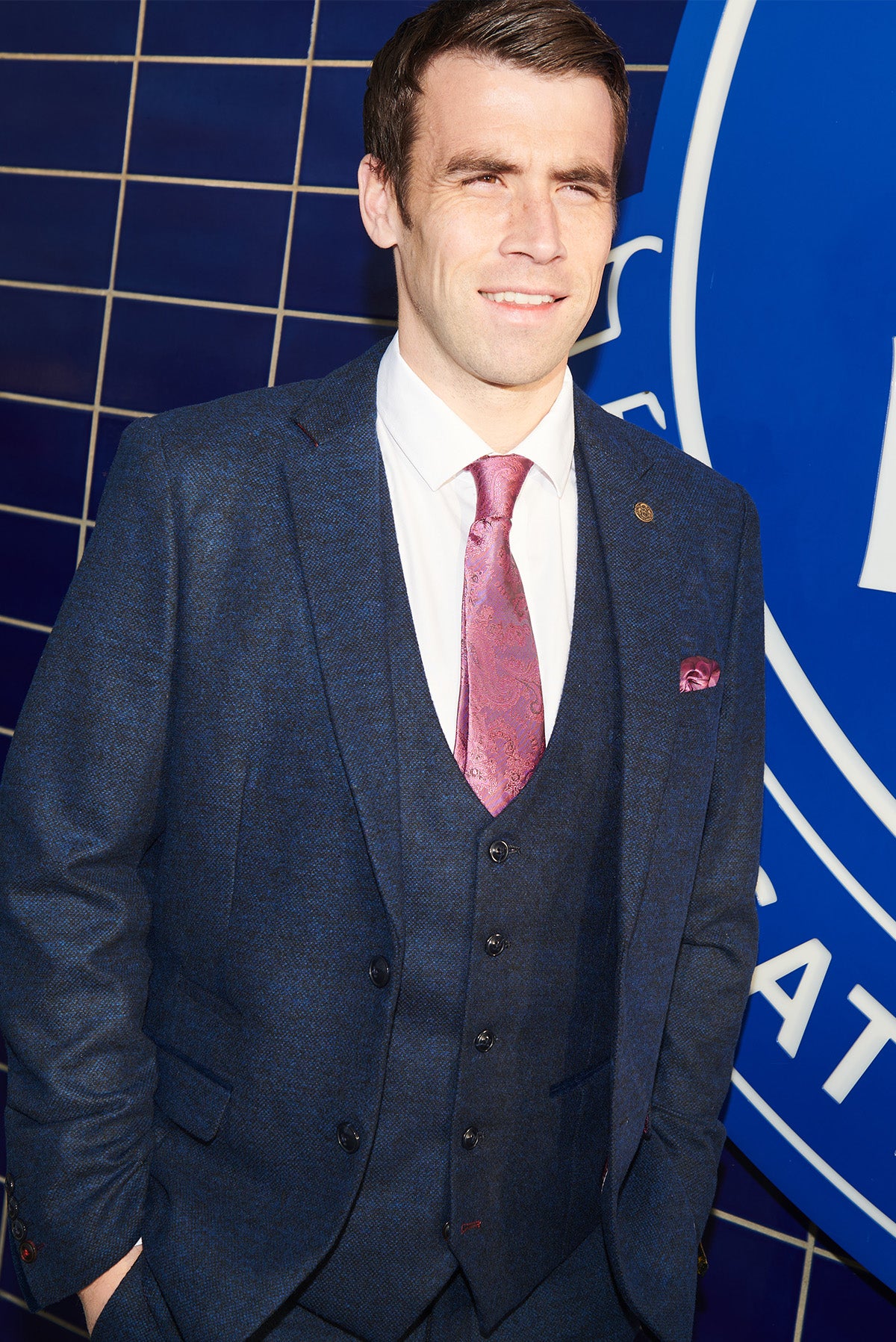 Séamus Coleman smiles in the MARLOW Navy Tweed Suit from marcdarcy’s Everton Collection, styled with a white shirt and patterned pink tie, standing against a blue tiled wall and circular sign.