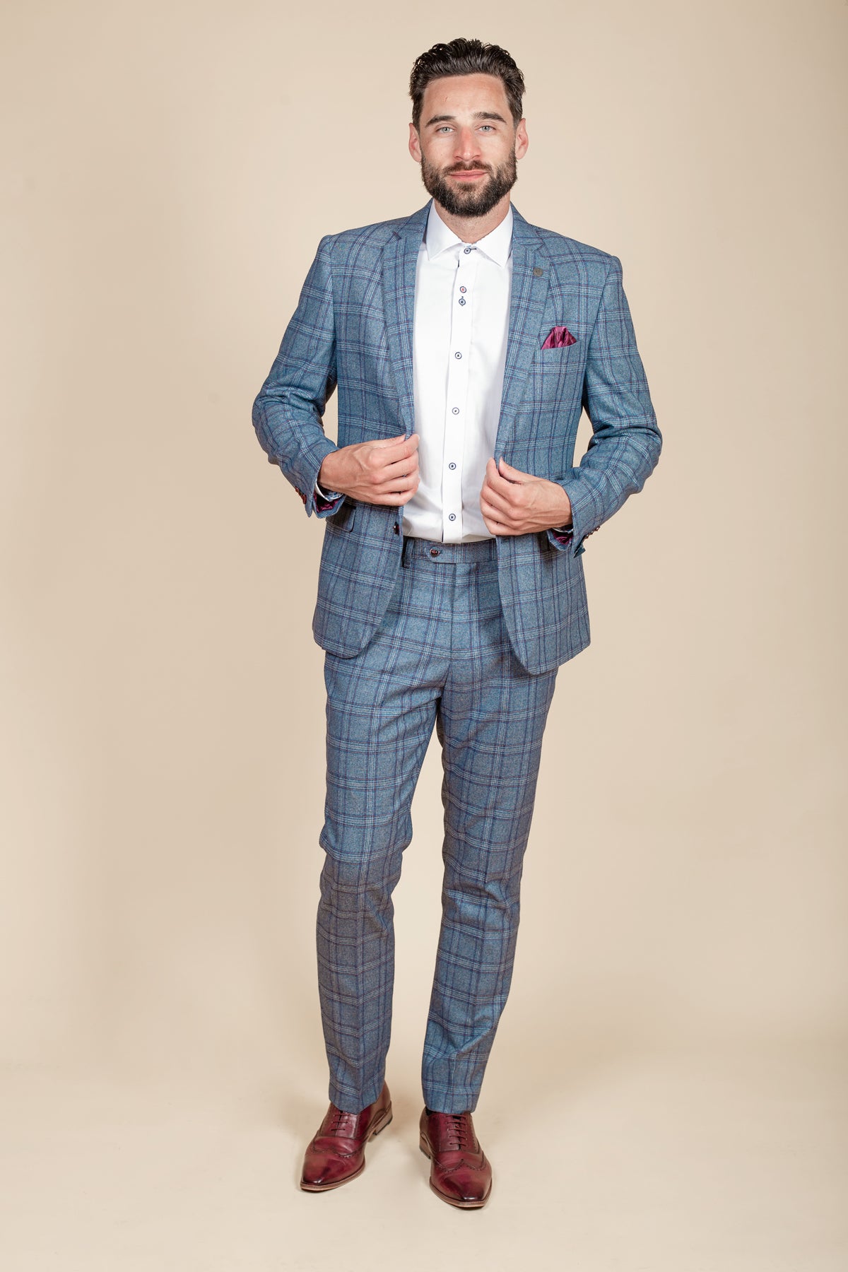 A man with dark hair and a beard stands facing forward, wearing the marcdarcy ABBOTT Blue Tweed Check Blazer over a white dress shirt and brown dress shoes, holding the sides of his open jacket against a beige background.