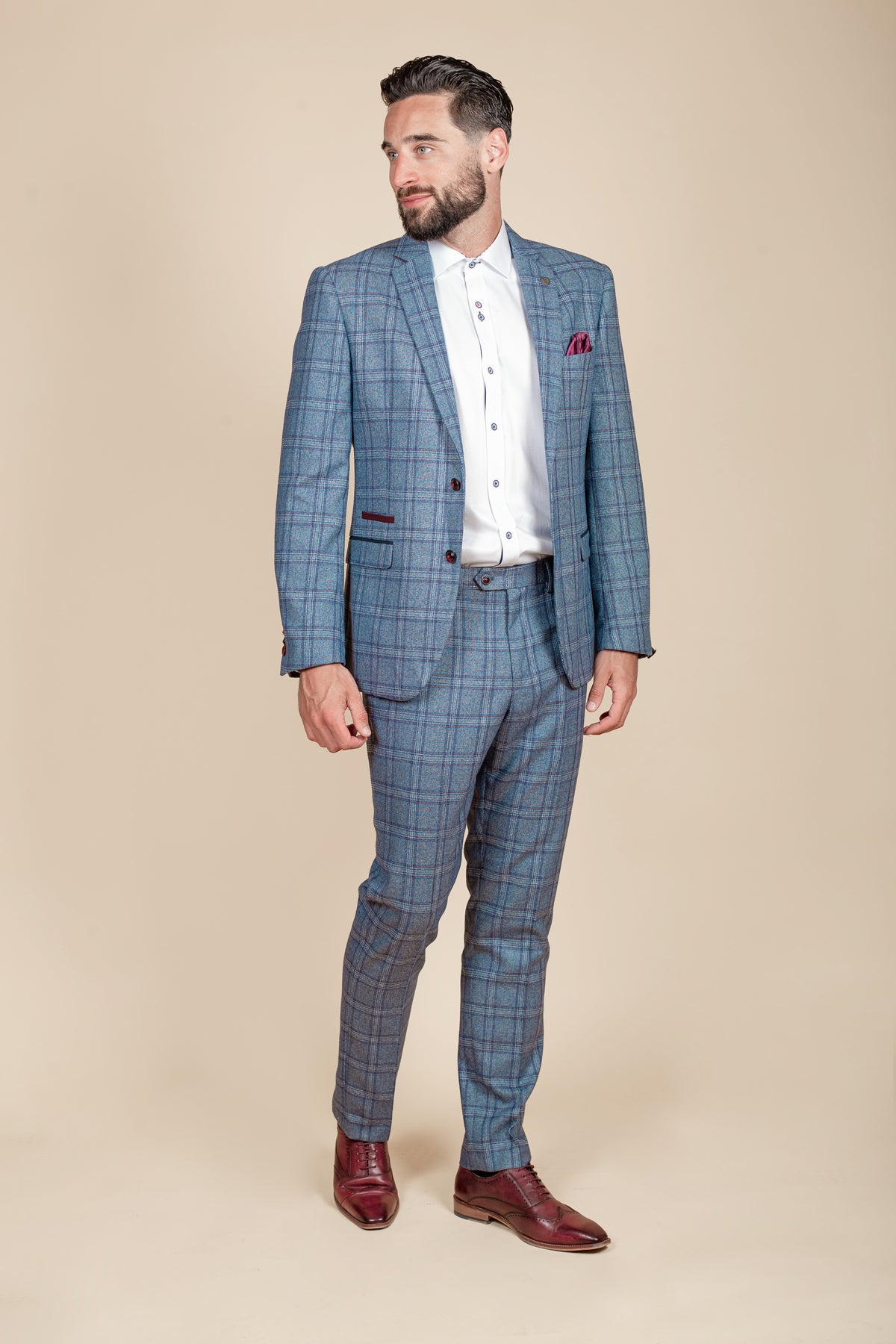 A man in the marcdarcy ABBOTT Blue Tweed Check Blazer stands on a beige background wearing a white shirt, brown dress shoes, and no tie. He embodies contemporary heritage style with short dark hair, a beard, and looks to his right.