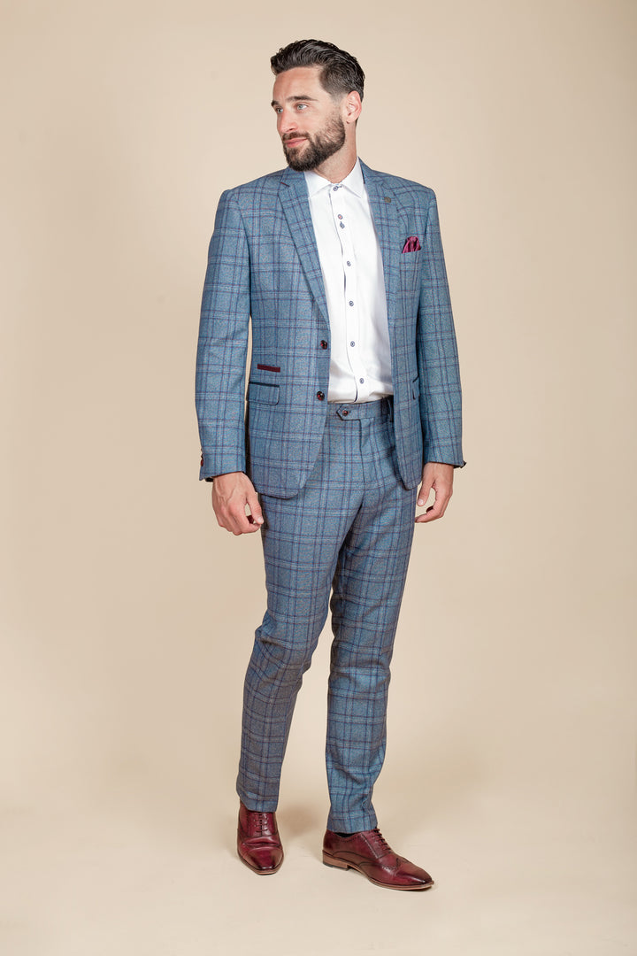 A man with short dark hair and a beard wears the marcdarcy ABBOTT Blue Tweed Check Blazer, a white dress shirt, and burgundy dress shoes. He stands against a beige background, glancing left with one hand relaxed at his side.