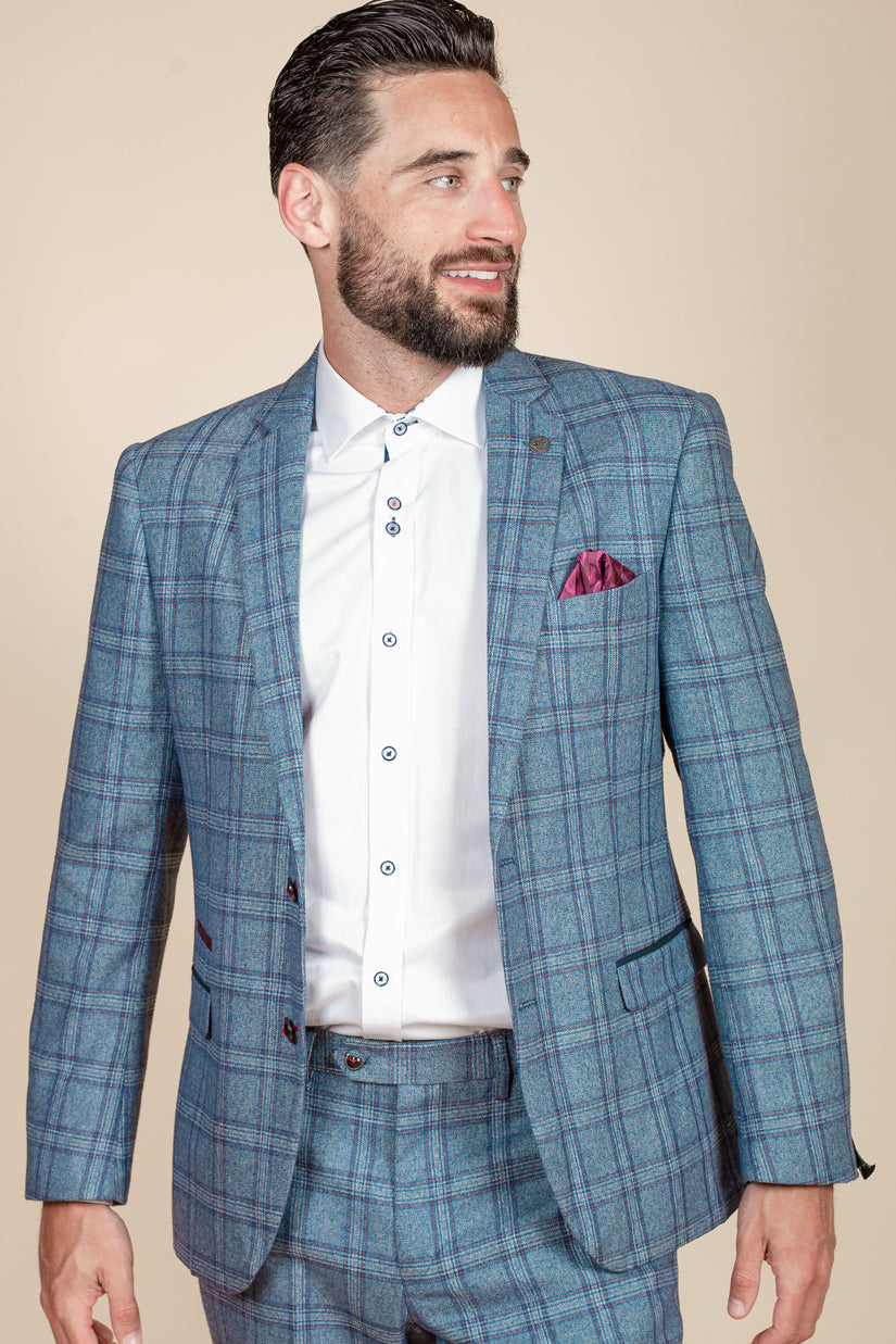 A man with dark hair and a beard wears the marcdarcy ABBOTT Blue Tweed Check Blazer with a white shirt and pink pocket square, standing against a beige background and looking to his left.